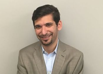 Samuel Breier wearing a light blue shirt and tan blazer, smiles against a plain light gray wall.