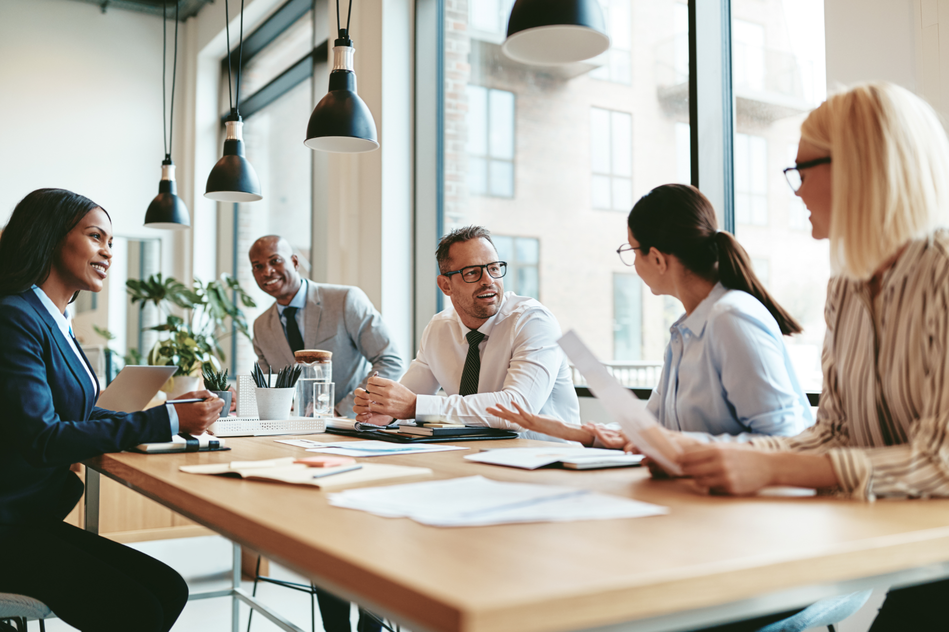 A diverse group of professionals in business attire collaborating around a table in a bright, modern office conference room.