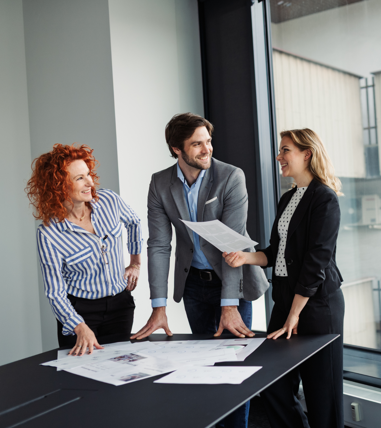 Three colleagues collaborate over architectural blueprints at a modern office table, smiling and discussing a document.