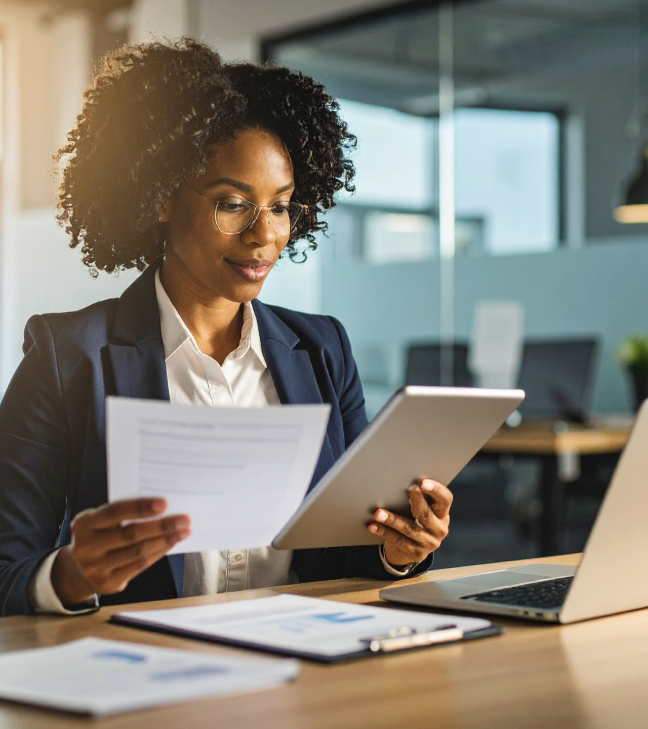 A professional works at a desk, reviewing a document and tablet while a laptop sits nearby in a bright office.