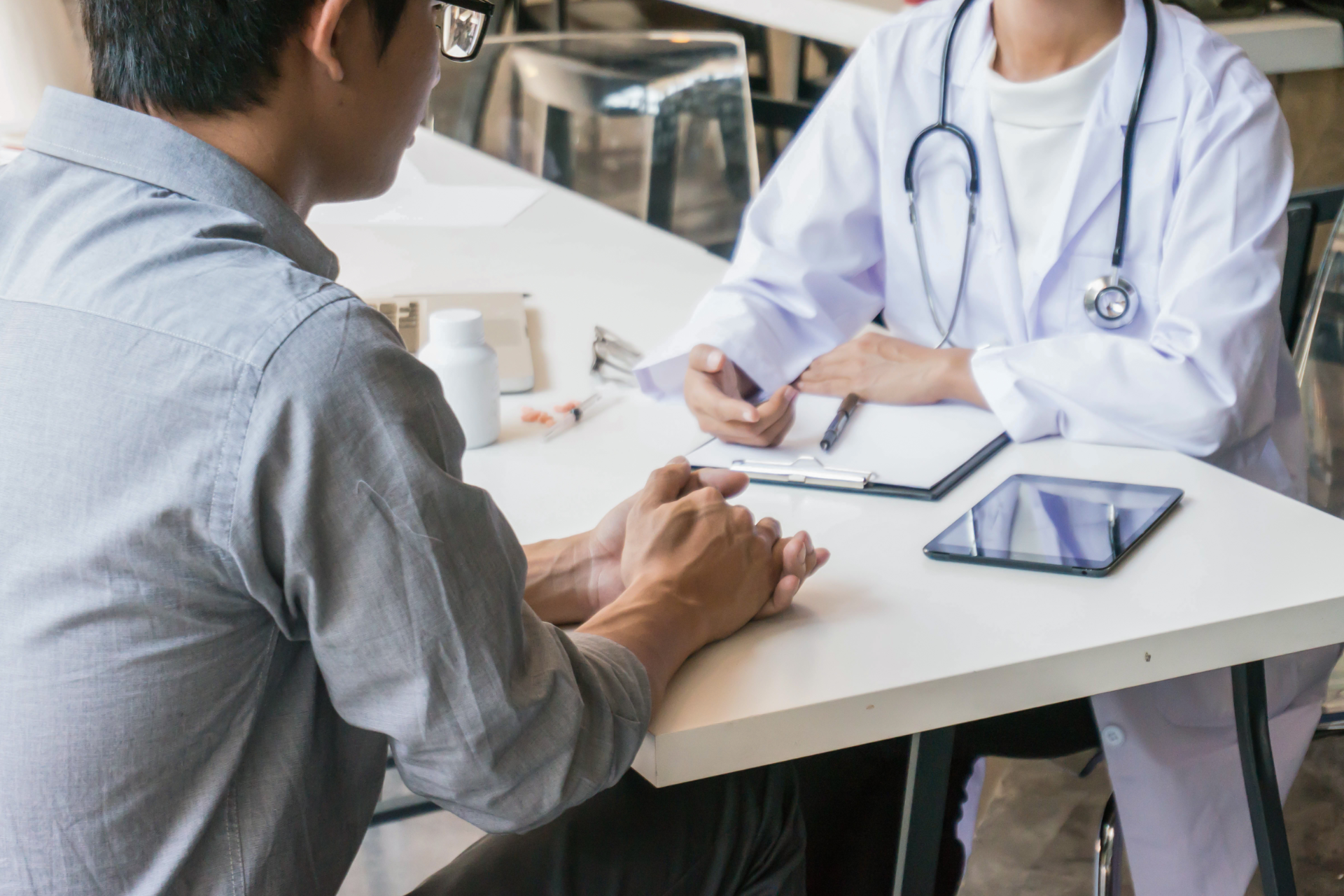 A patient sits across from a doctor in a white coat at a table, discussing medical information with a clipboard.