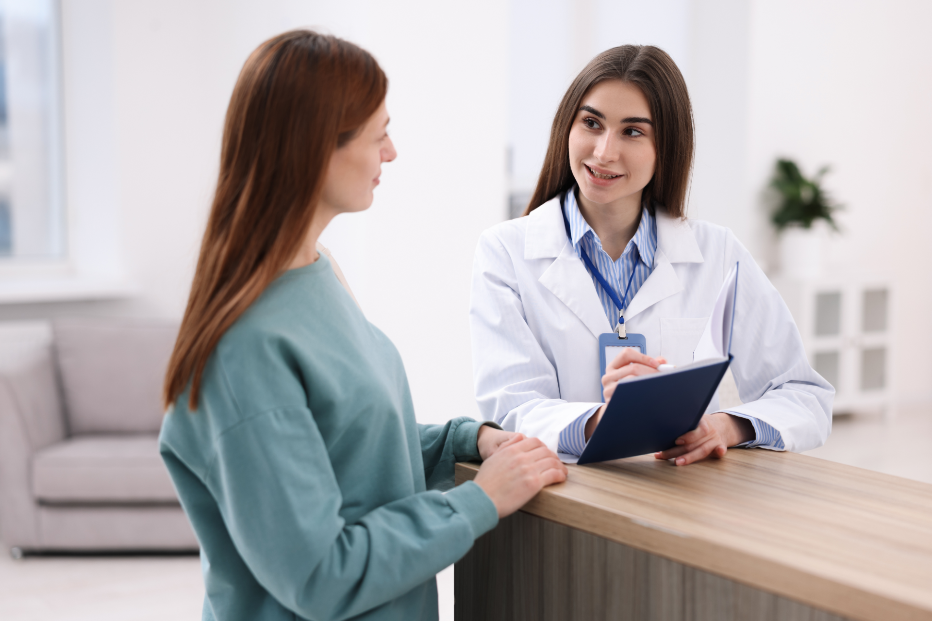 A person in a medical coat stands behind a counter, speaking with a client and holding a notepad in a bright office.