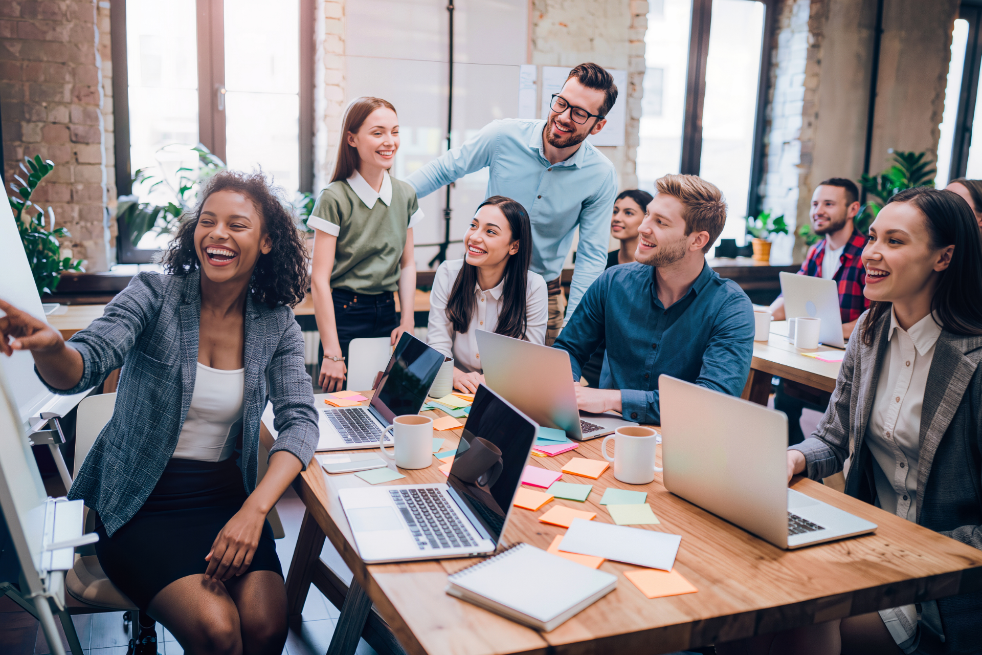 A diverse group of colleagues collaborate in a bright office, laughing and working together at a wooden table with laptops.