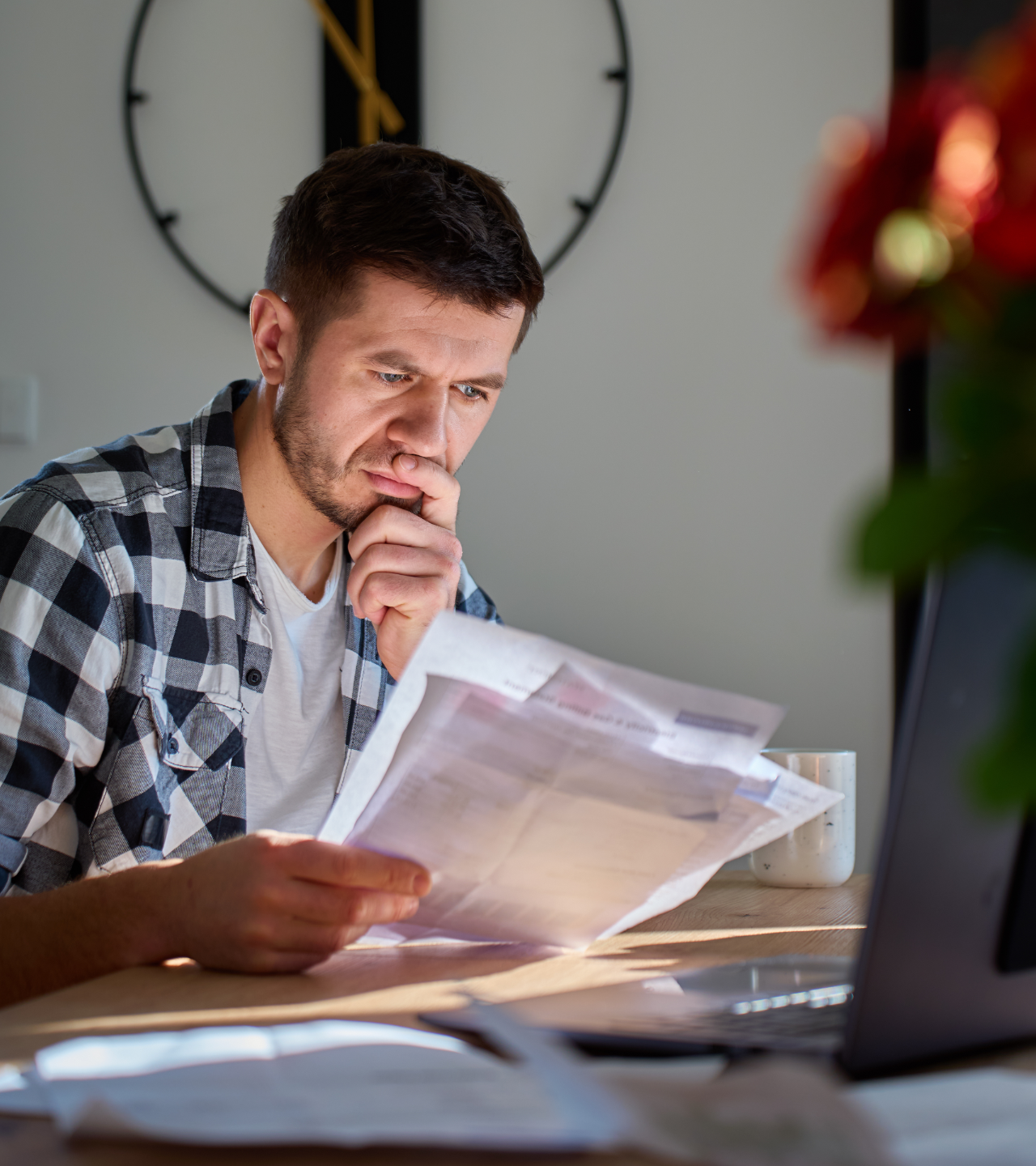 A man sits at a desk in a home office, looking at documents with a focused, thoughtful expression.