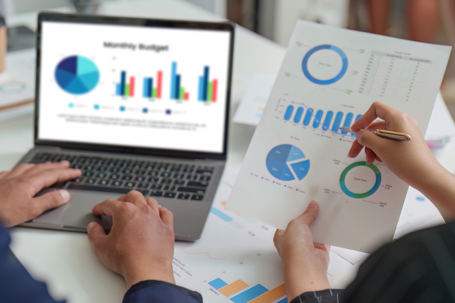 Two people analyze financial charts displayed on a laptop screen and a printed document in a professional office setting.