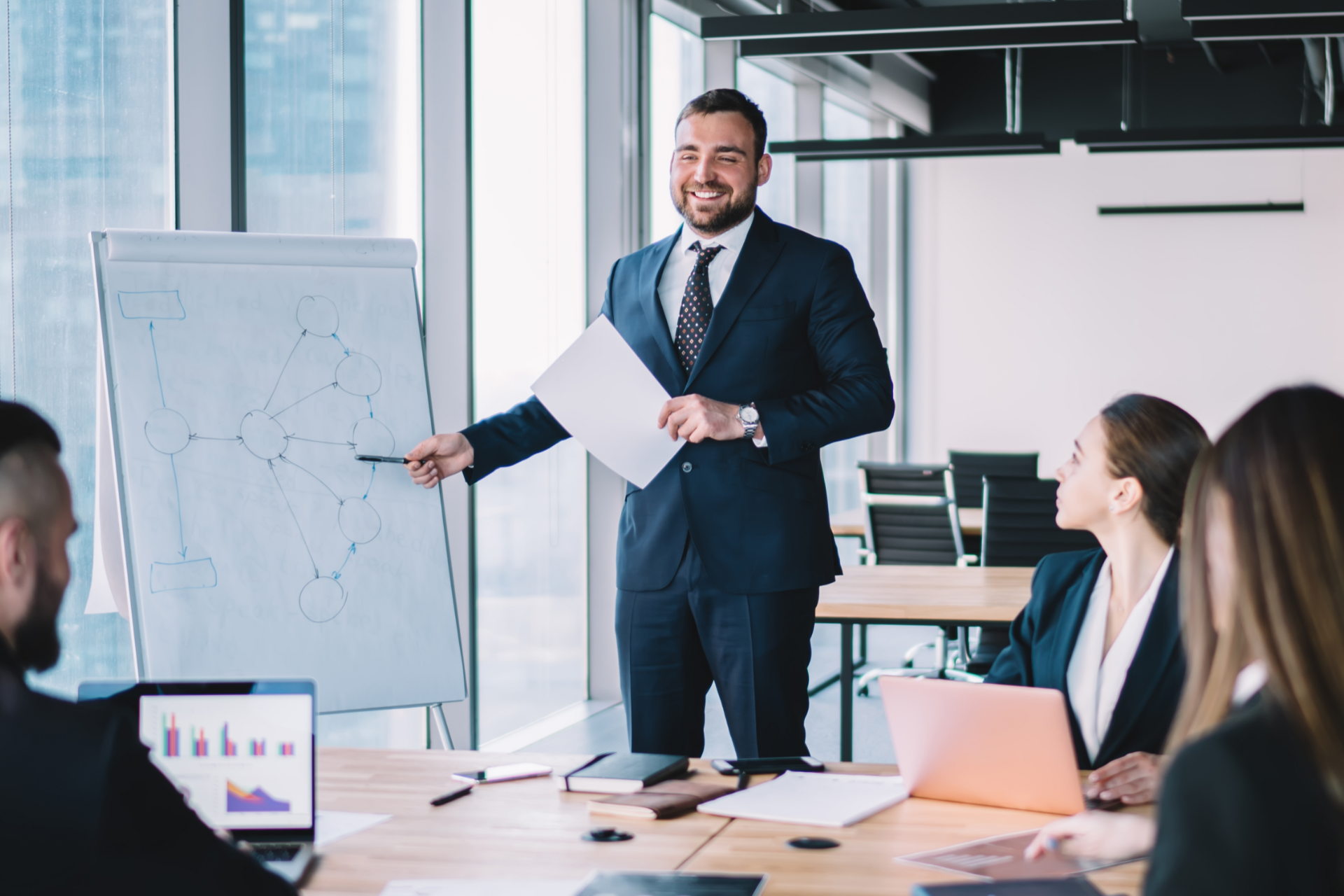 A professional leads a meeting in a bright office, pointing to a diagram on a whiteboard with colleagues watching.