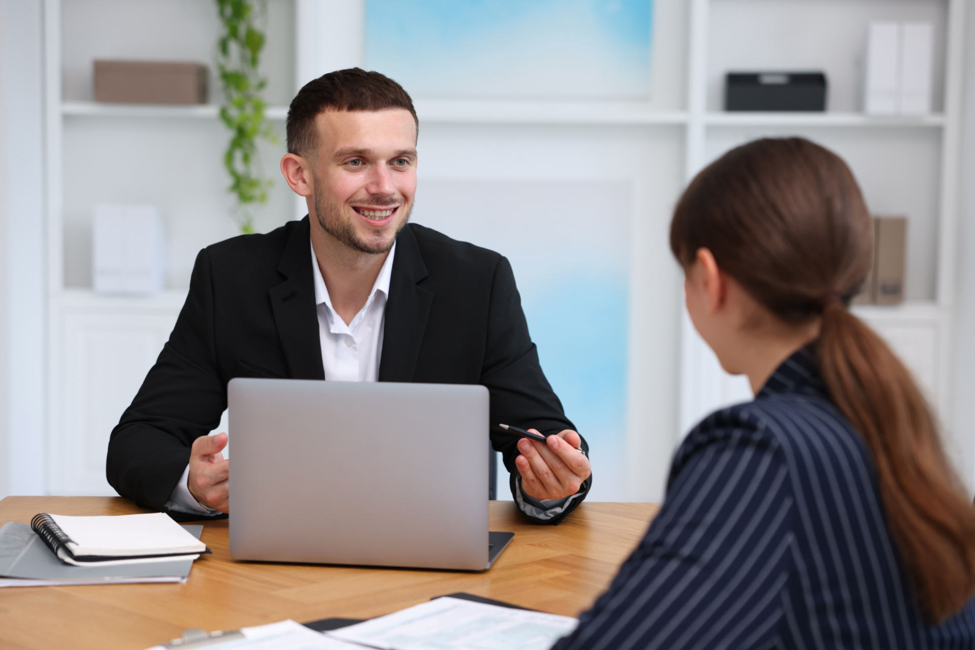 A smiling professional in a suit gestures while speaking to a colleague across a desk in a bright office.