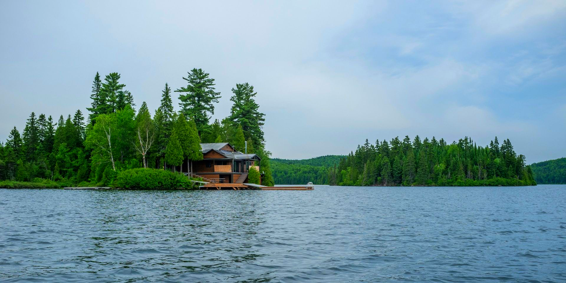 A wooden cabin sits on a small, tree-covered island in the middle of a large, rippling lake under a cloudy sky.