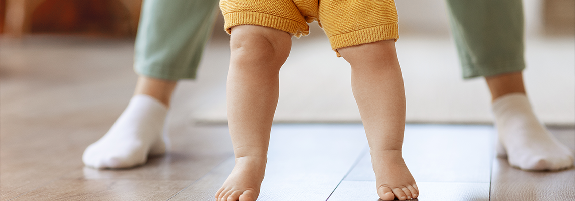 Baby learning to walk on a tiled floor, with adult legs supporting behind