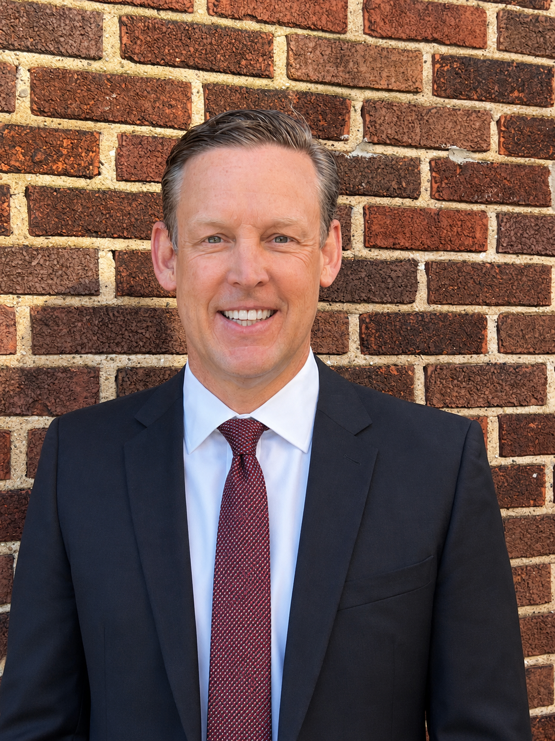 Man in a dark suit and red tie smiling in front of a brick wall