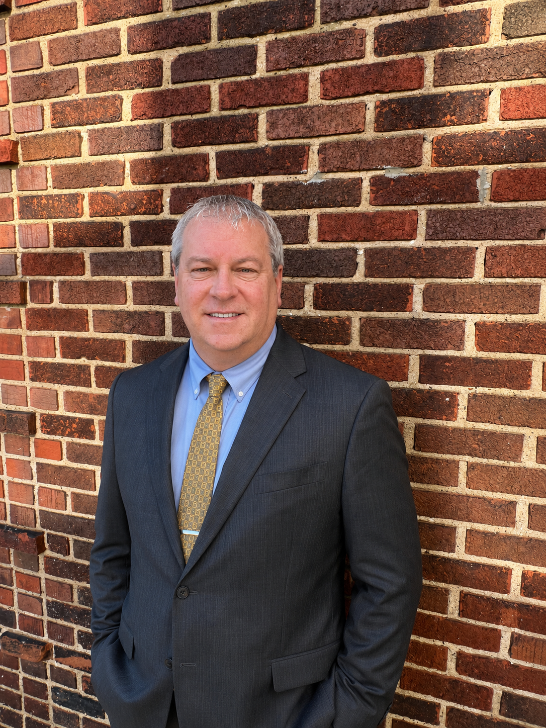 Man in a dark suit and gold tie standing against a brick wall, smiling at the camera