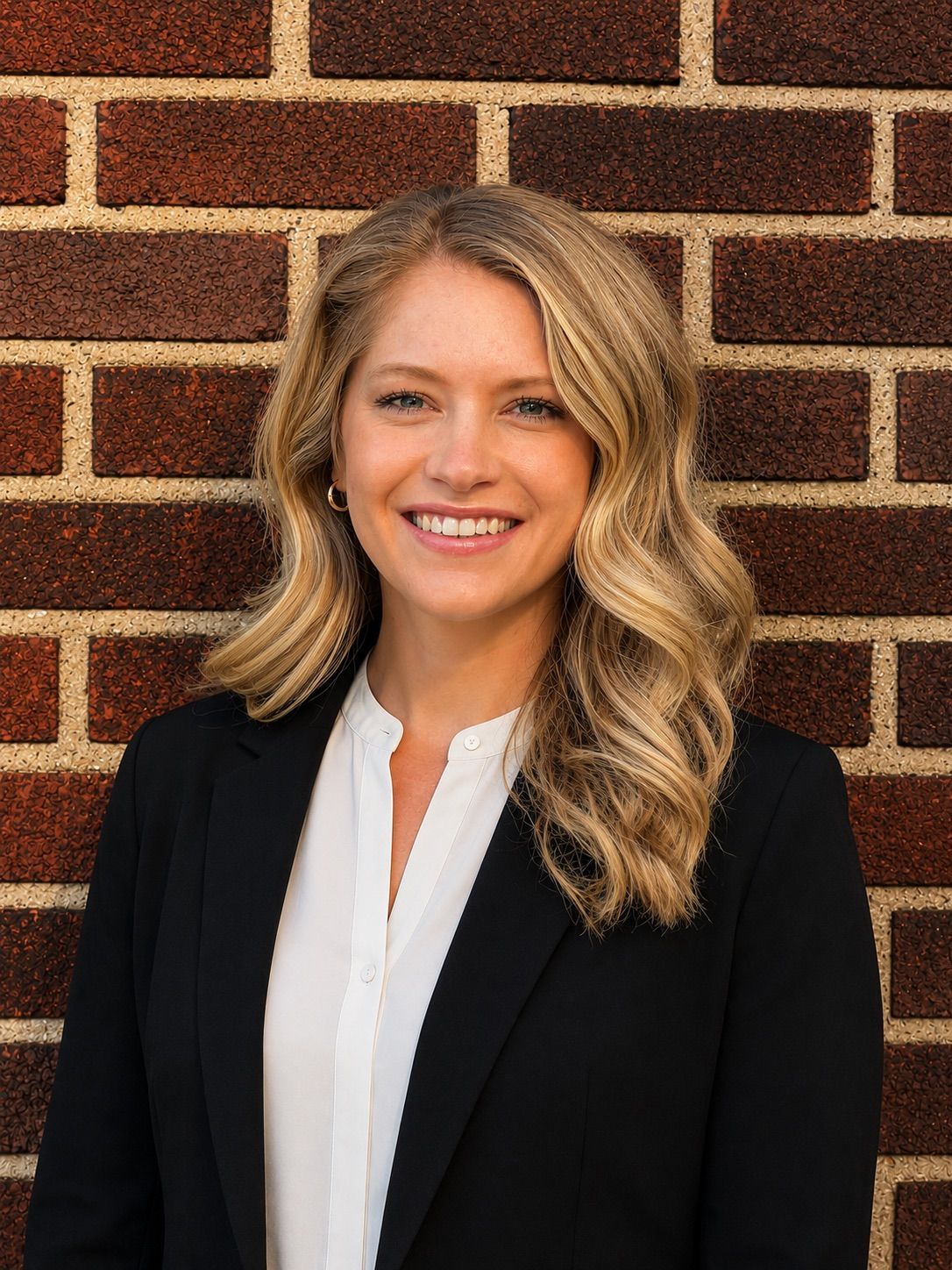 Smiling woman in a black blazer and white blouse standing against a brick wall