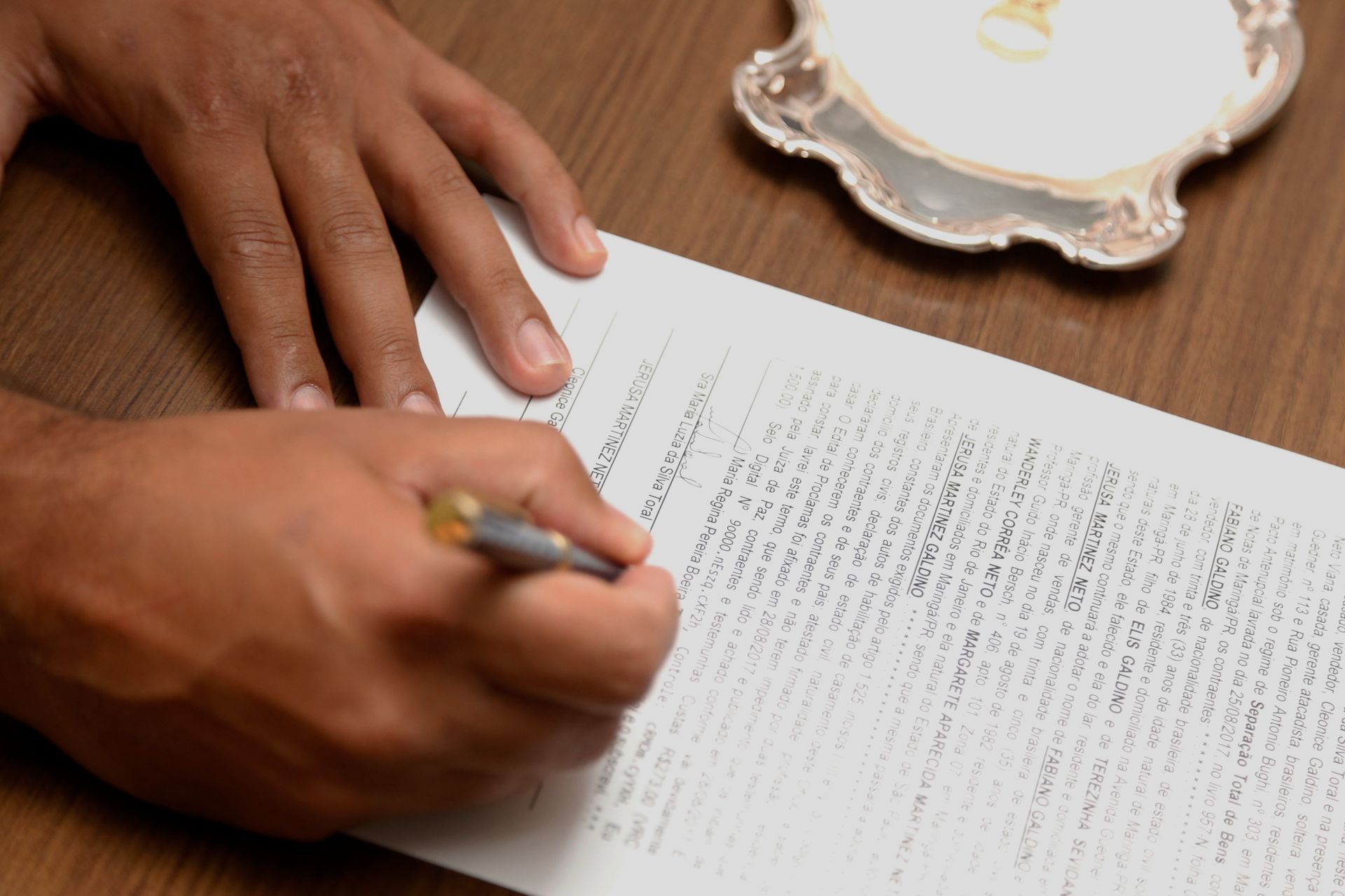 Hands signing a document at a wooden table beside a small plate.