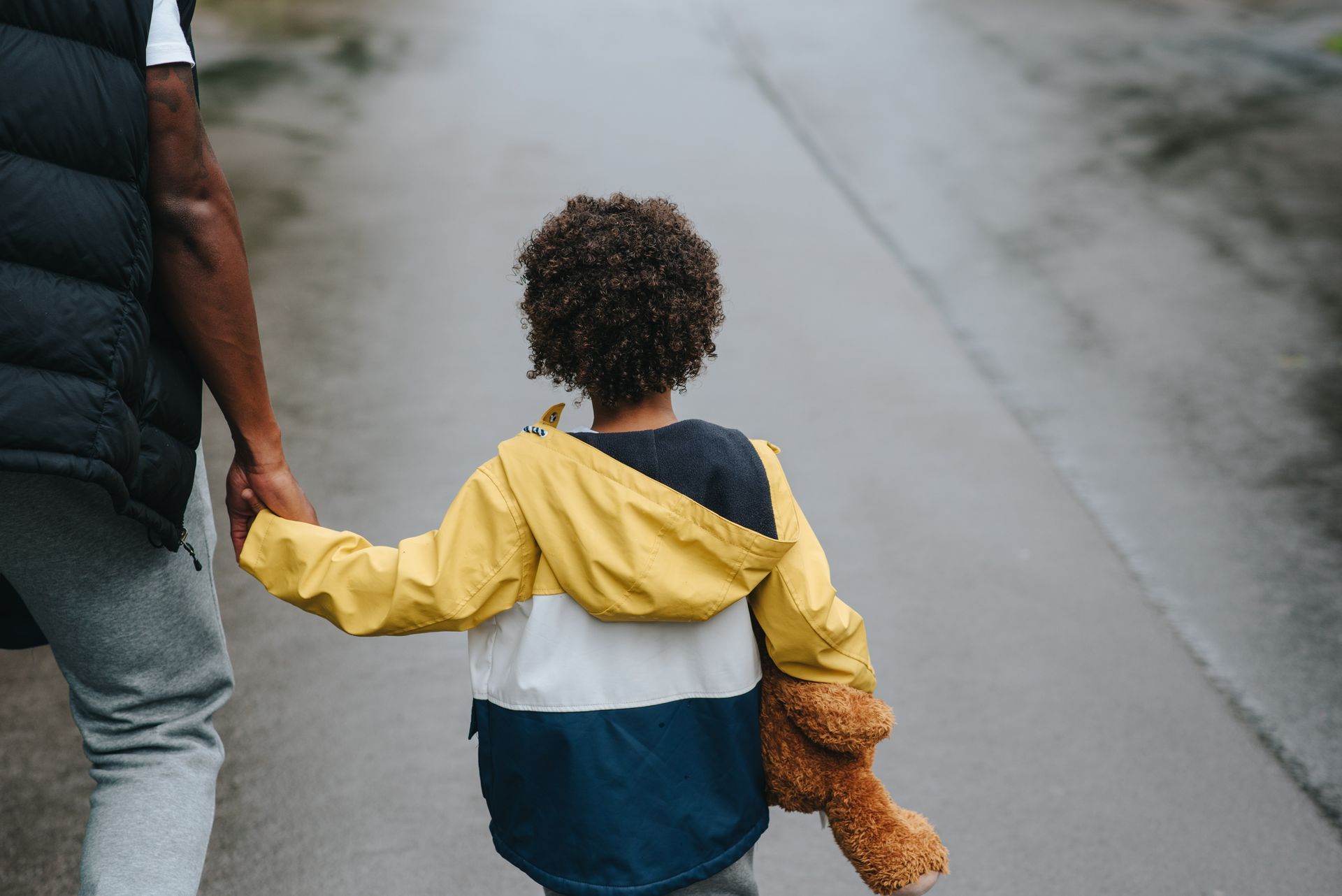 Adult and child holding hands, walking down a wet street with a teddy bear in the child’s other hand.