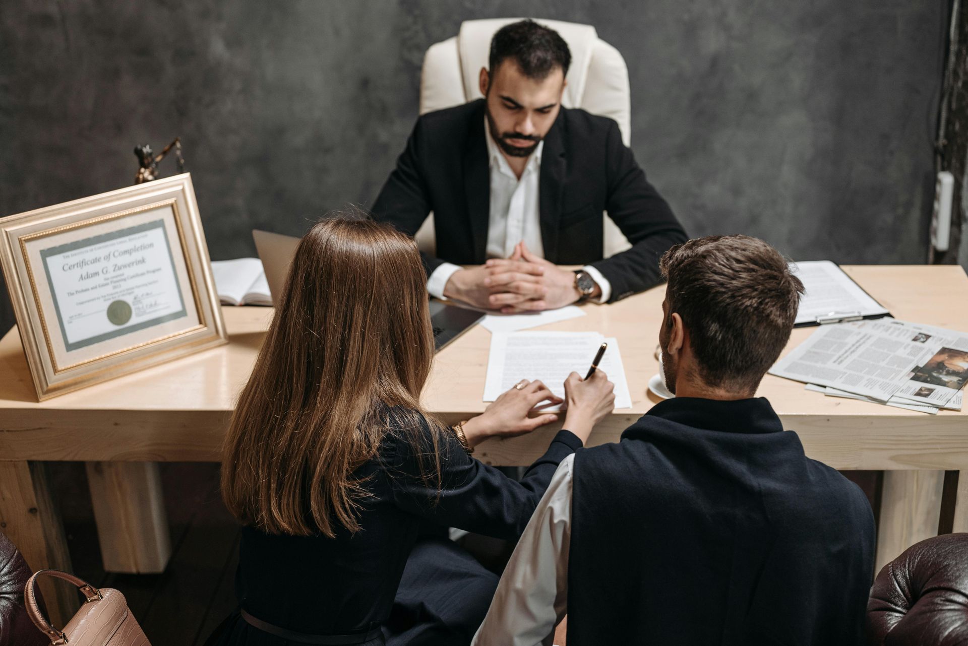 Two people meet with a man in a suit at a desk, reviewing documents in an office.