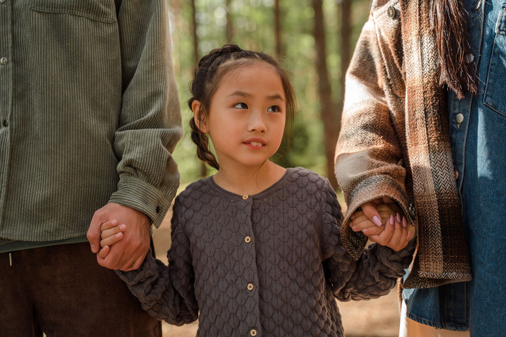 Three people holding hands outdoors in a wooded area, with a child in the center.