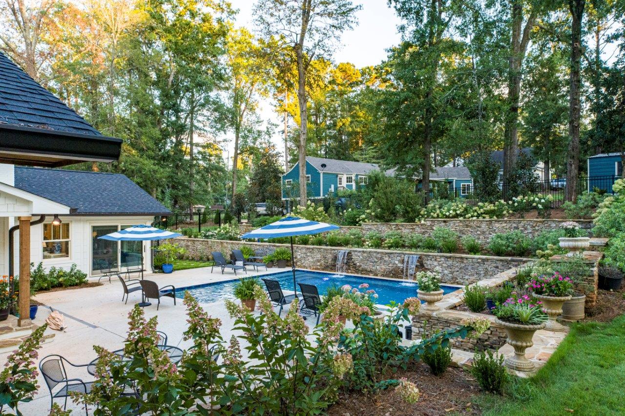 Backyard with pool, patio, stone retaining walls, lush greenery, and houses in background.