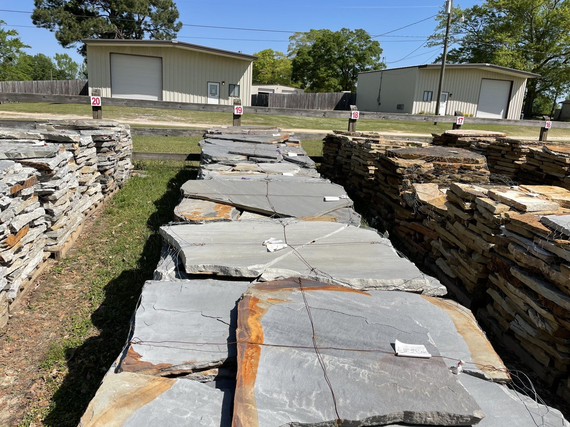 Stacks of flagstone pavers in a yard, with two industrial buildings visible in the background under a bright blue sky.