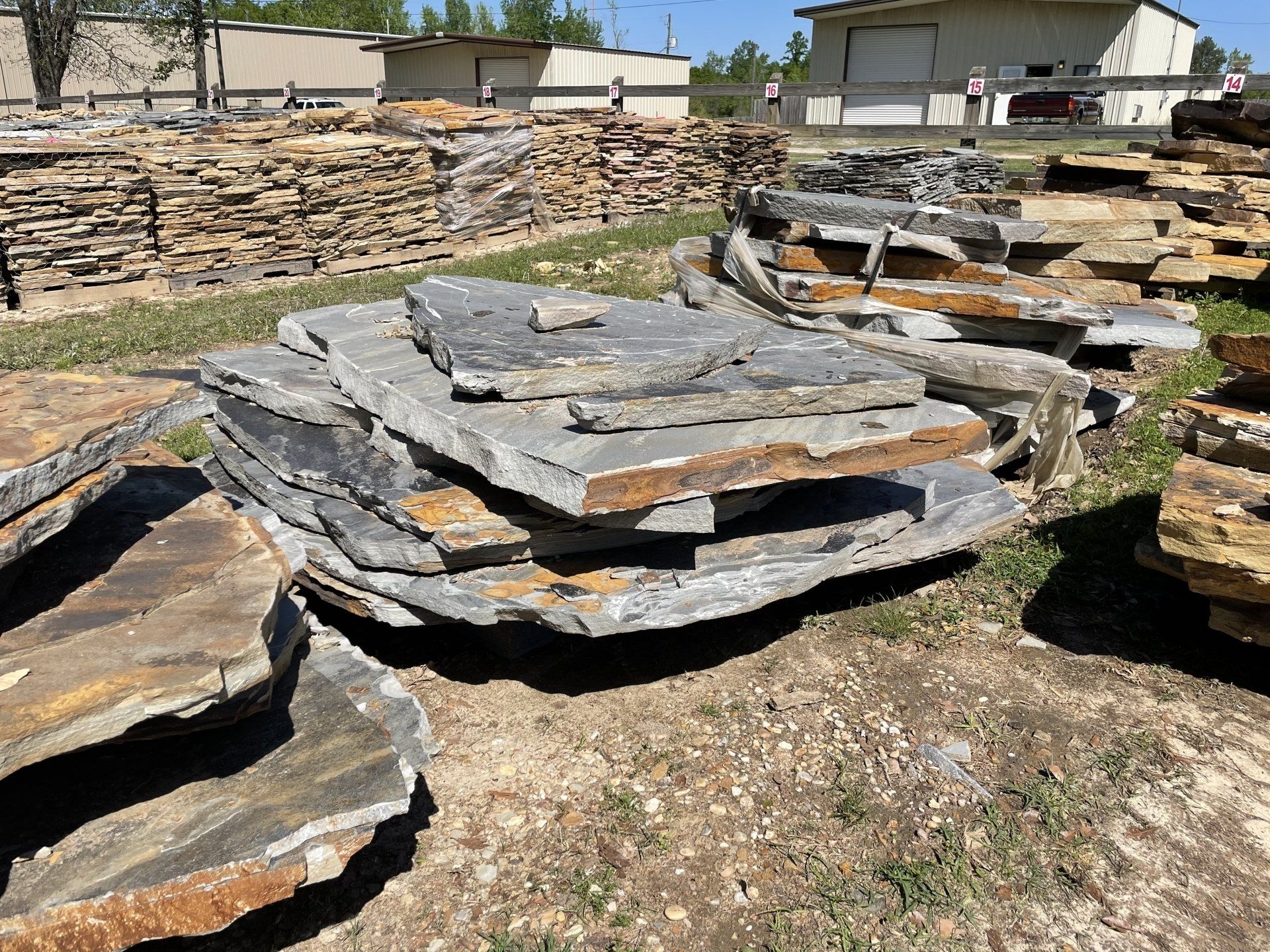 Pile of grey and brown flagstone slabs on grass, with more stone and a building in the background.