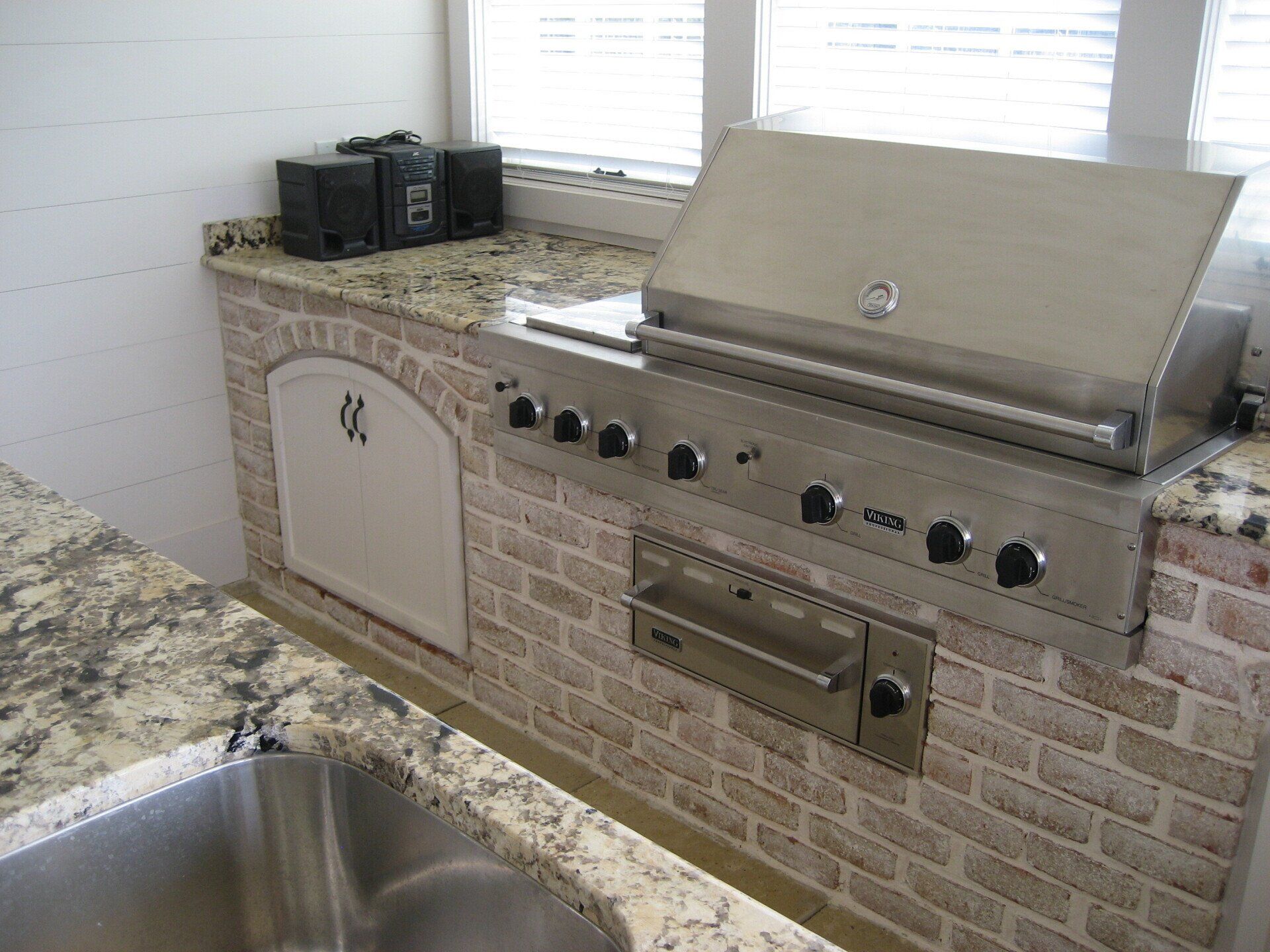 A stainless steel grill is built into a brick wall next to a sink.