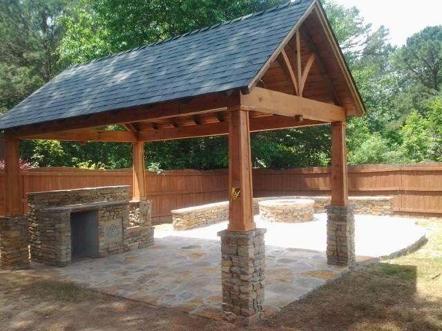 Wooden gazebo with stone fireplace and flooring, over a backyard fire pit area.