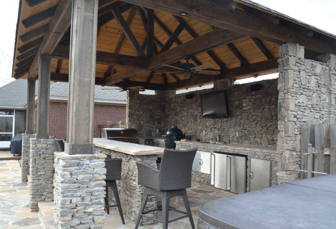 Outdoor kitchen with stone walls, wooden roof, bar, and seating.
