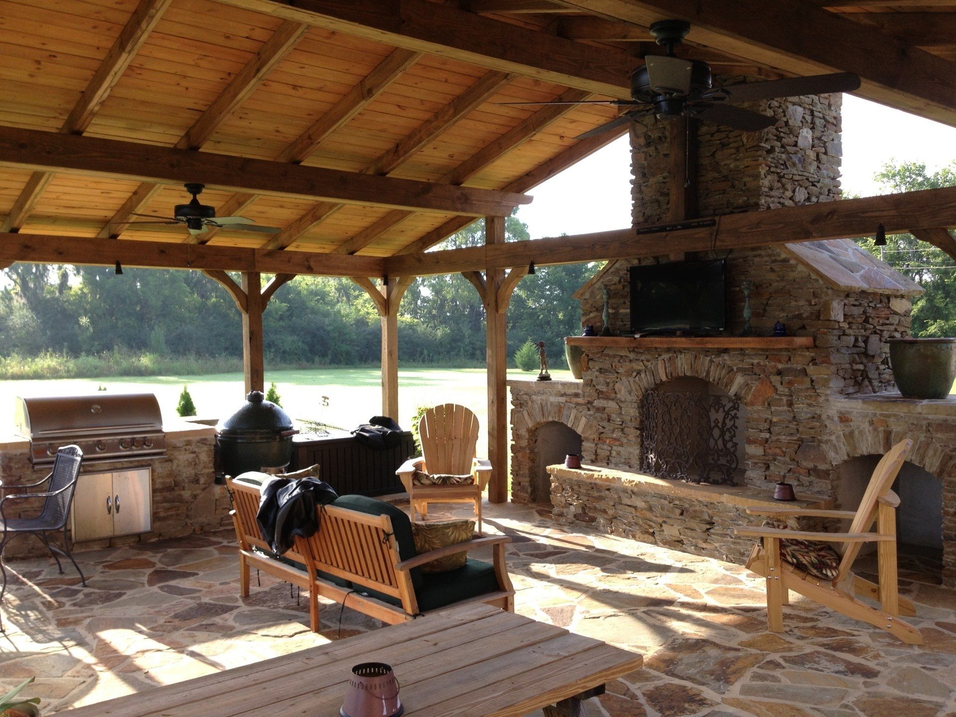 Covered outdoor kitchen with stone fireplace, grill, seating, and a view of a grassy field.