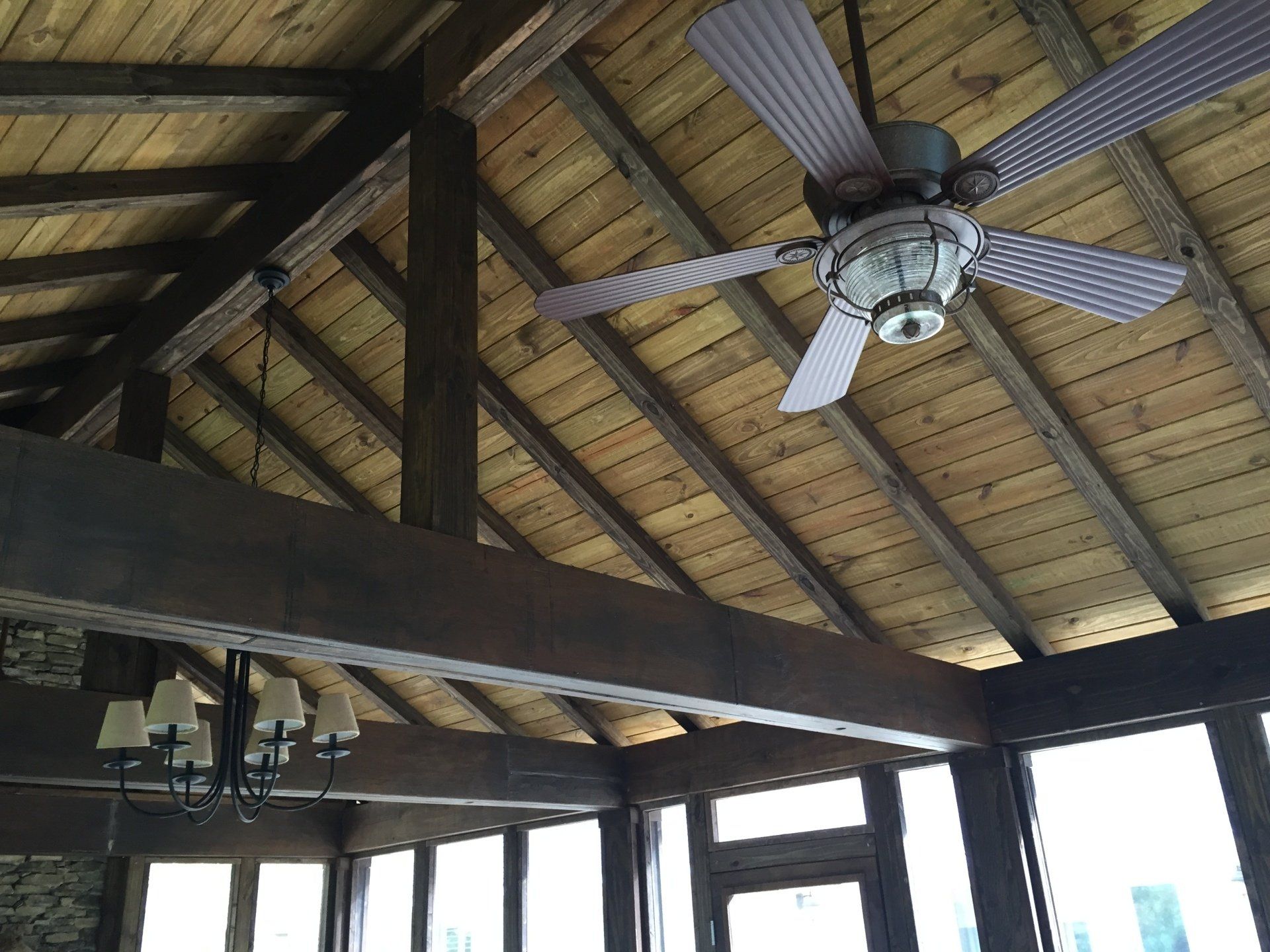Wooden ceiling with beams, a ceiling fan, and a chandelier. Large windows visible.