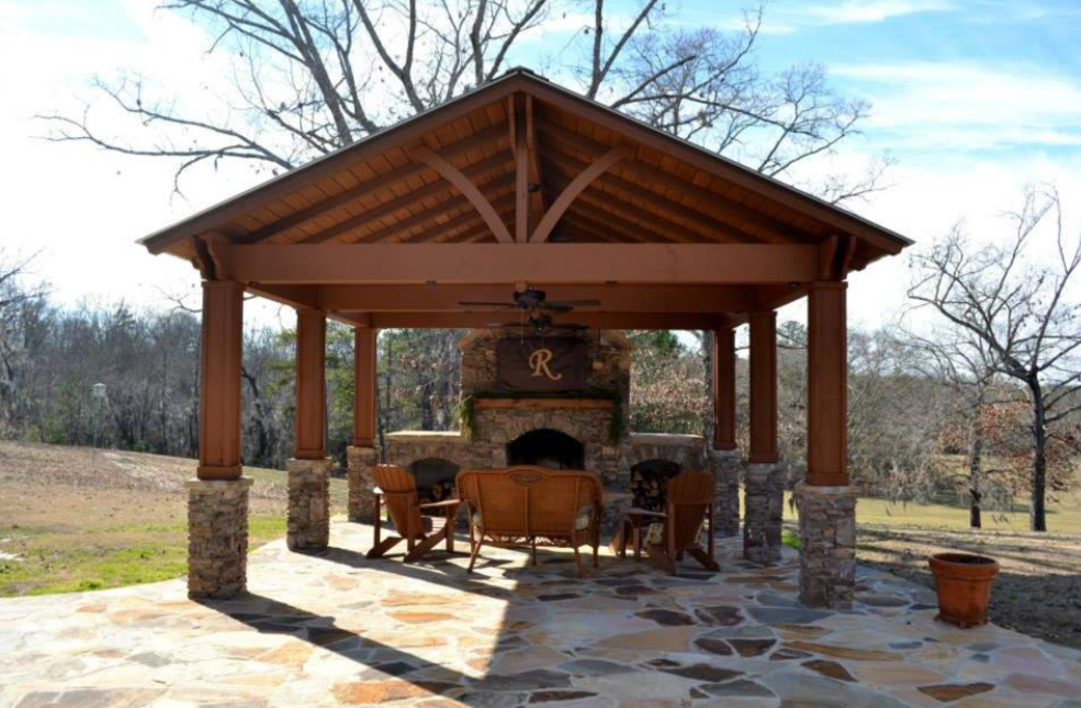 Outdoor pavilion with fireplace and seating on a stone patio. Wooden structure, stone accents, and surrounding trees.