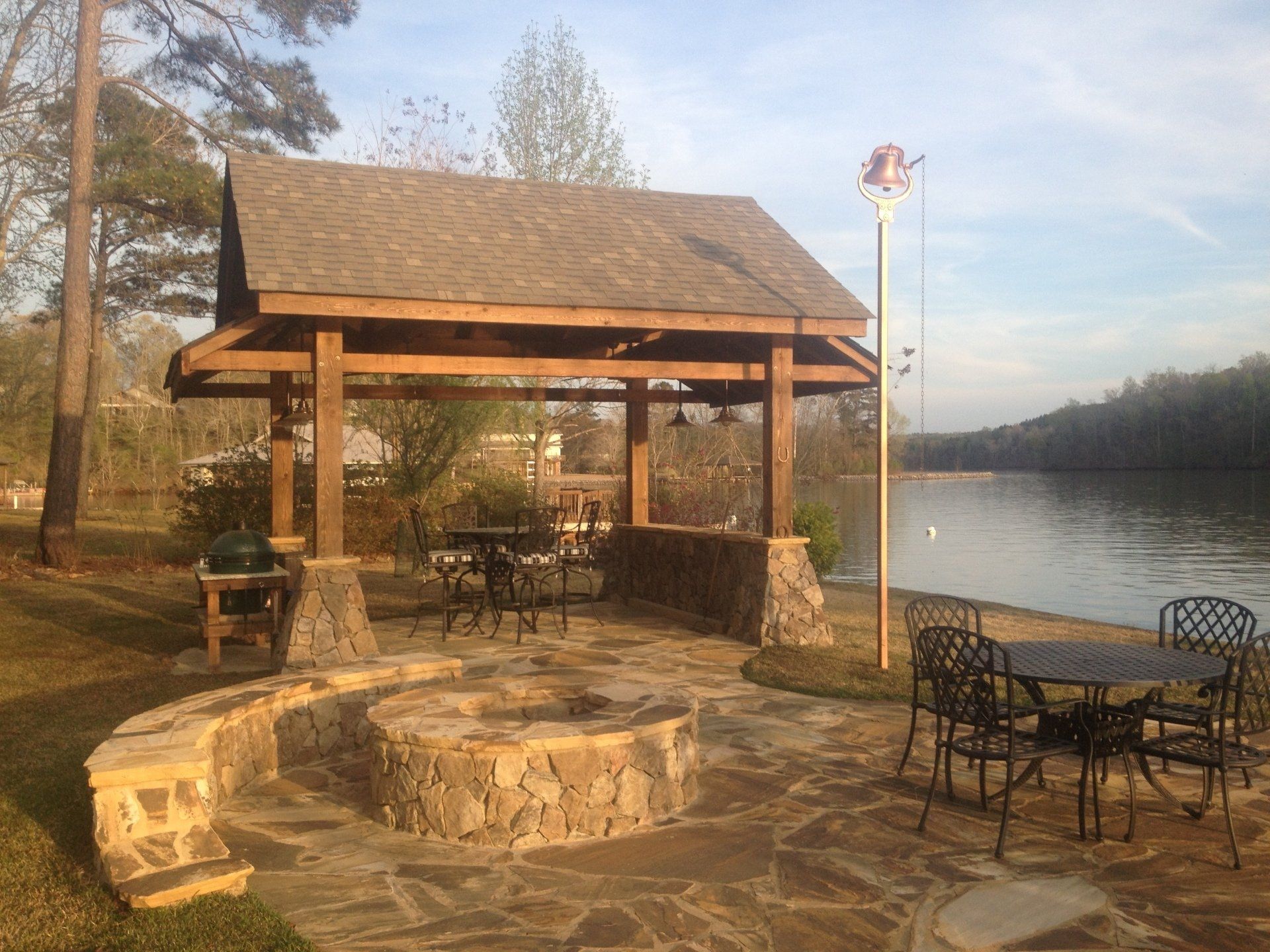 Patio with a stone fire pit, gazebo, and wrought-iron furniture overlooking a lake.