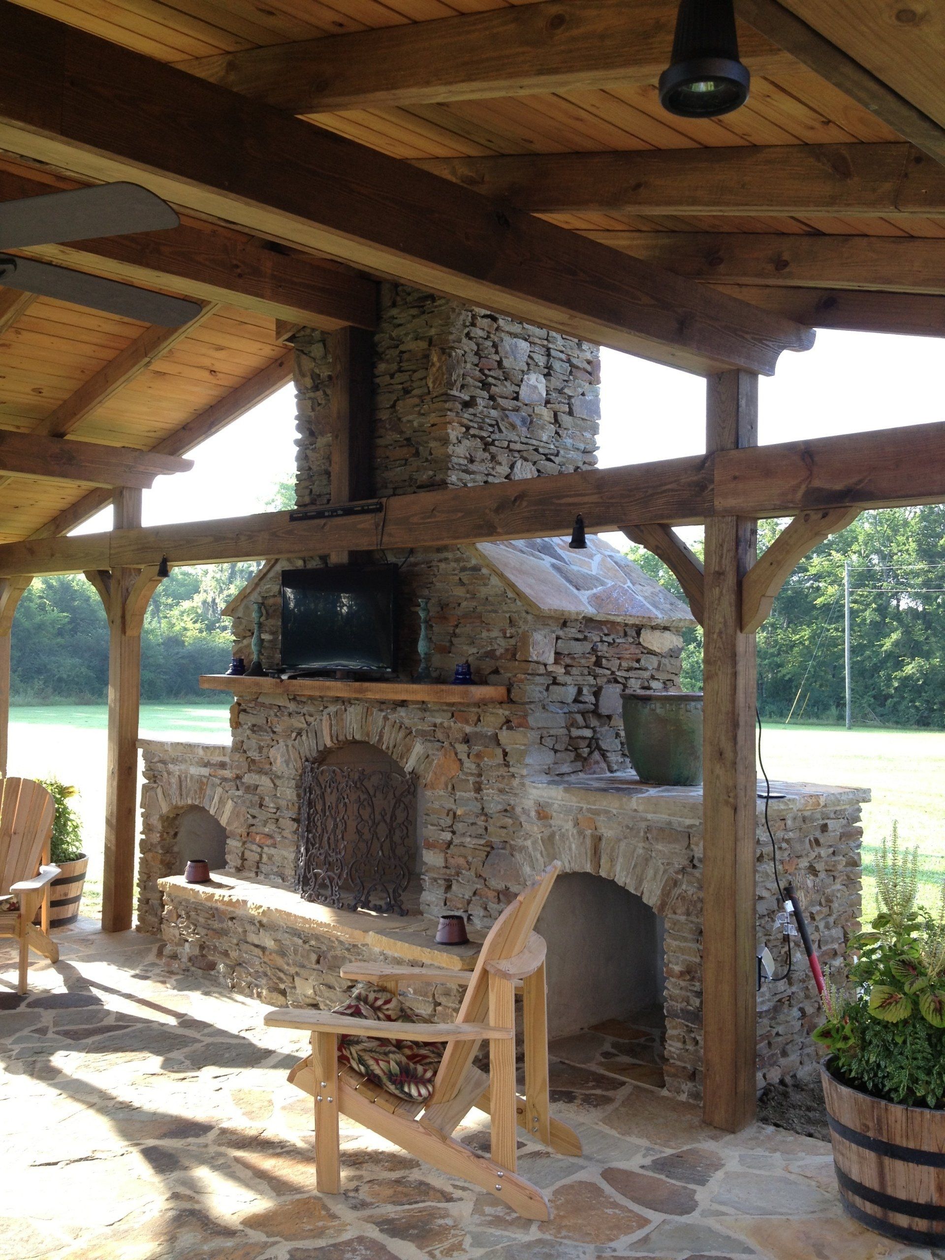 Stone fireplace with a TV, under a wooden pergola, next to a grassy field.