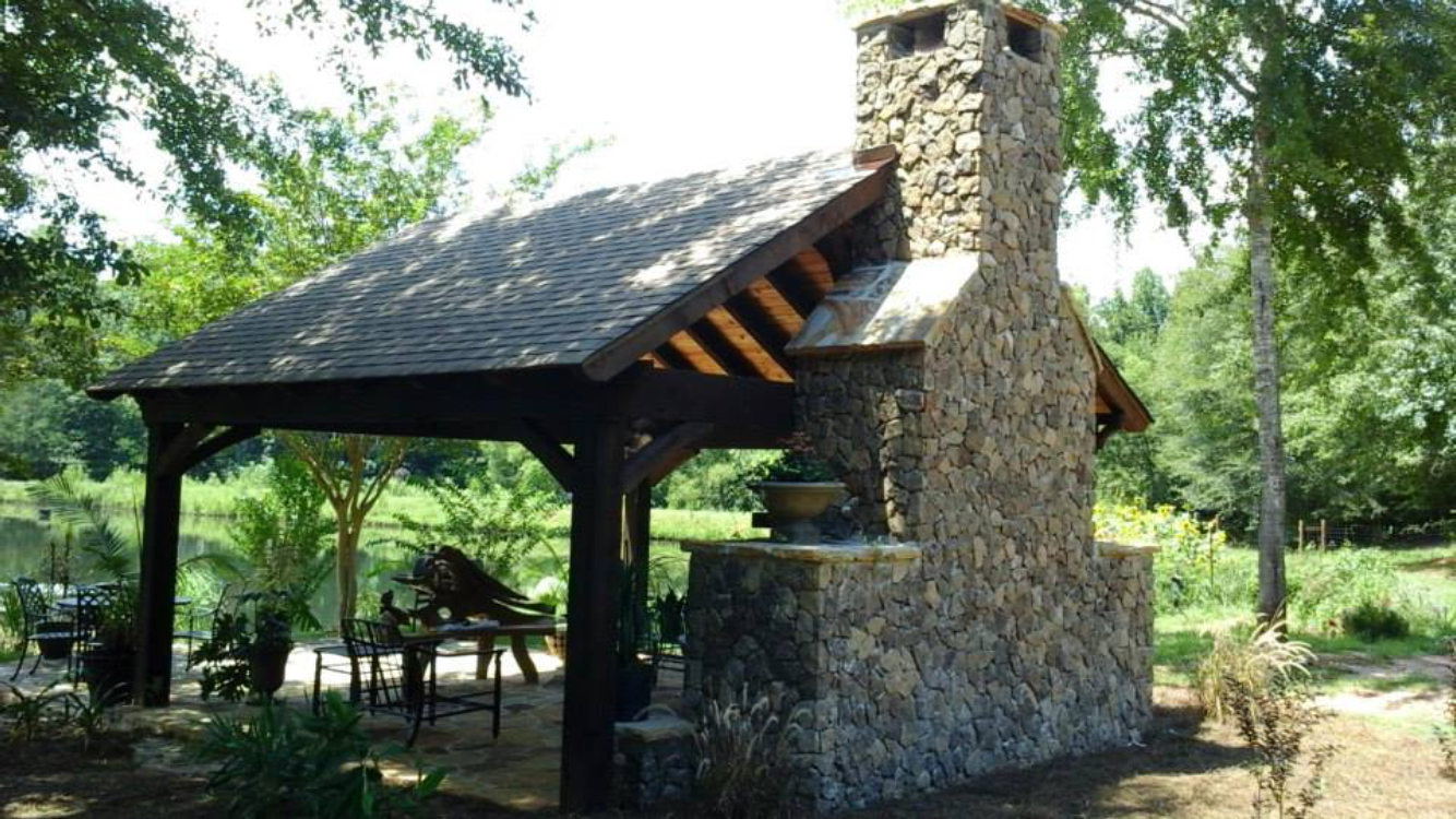 Outdoor stone fireplace with covered seating area. Brown roof and dark wood frame, natural surroundings.