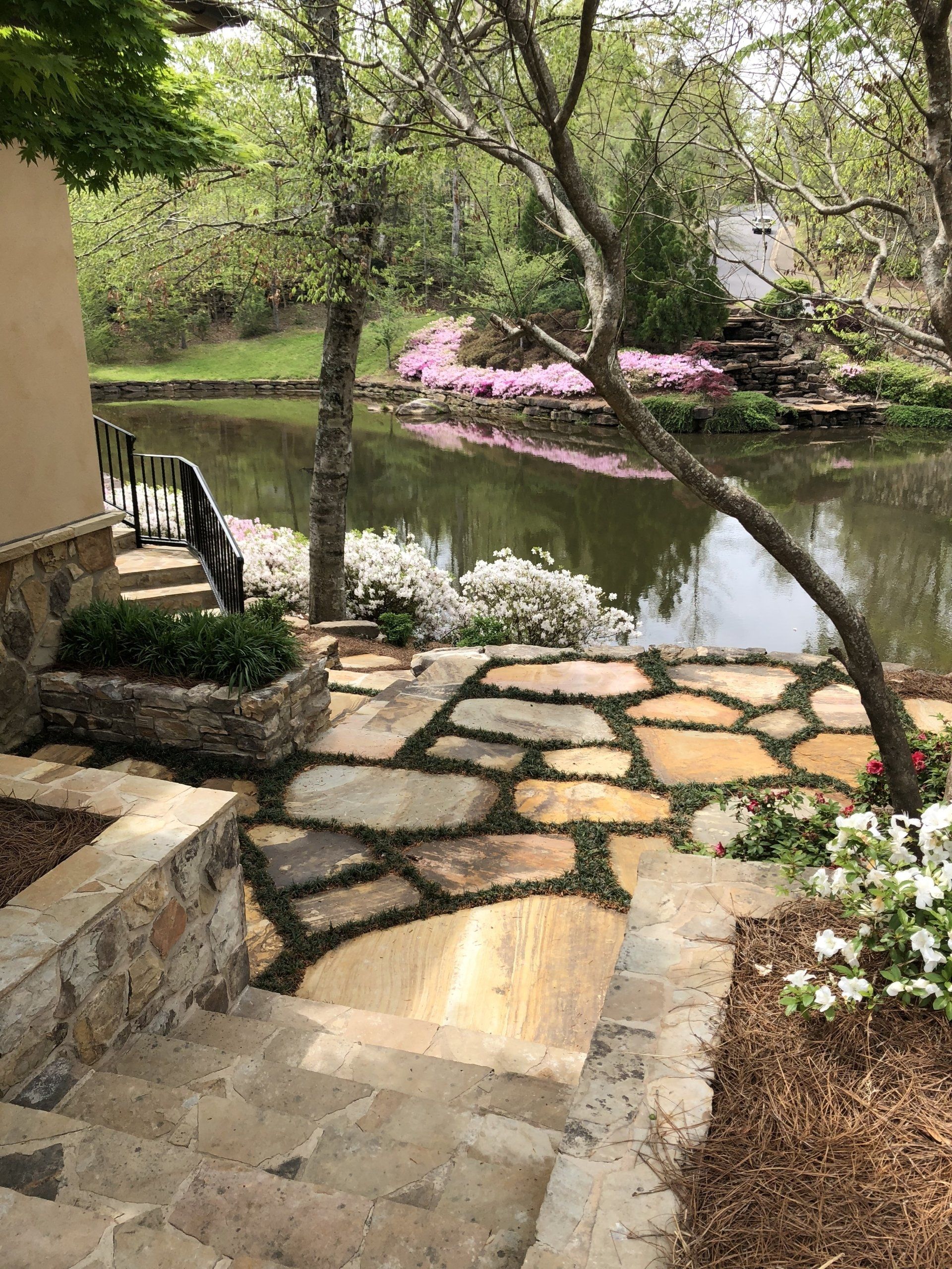 Stone patio and steps leading to a lake with blooming azaleas and a small waterfall.