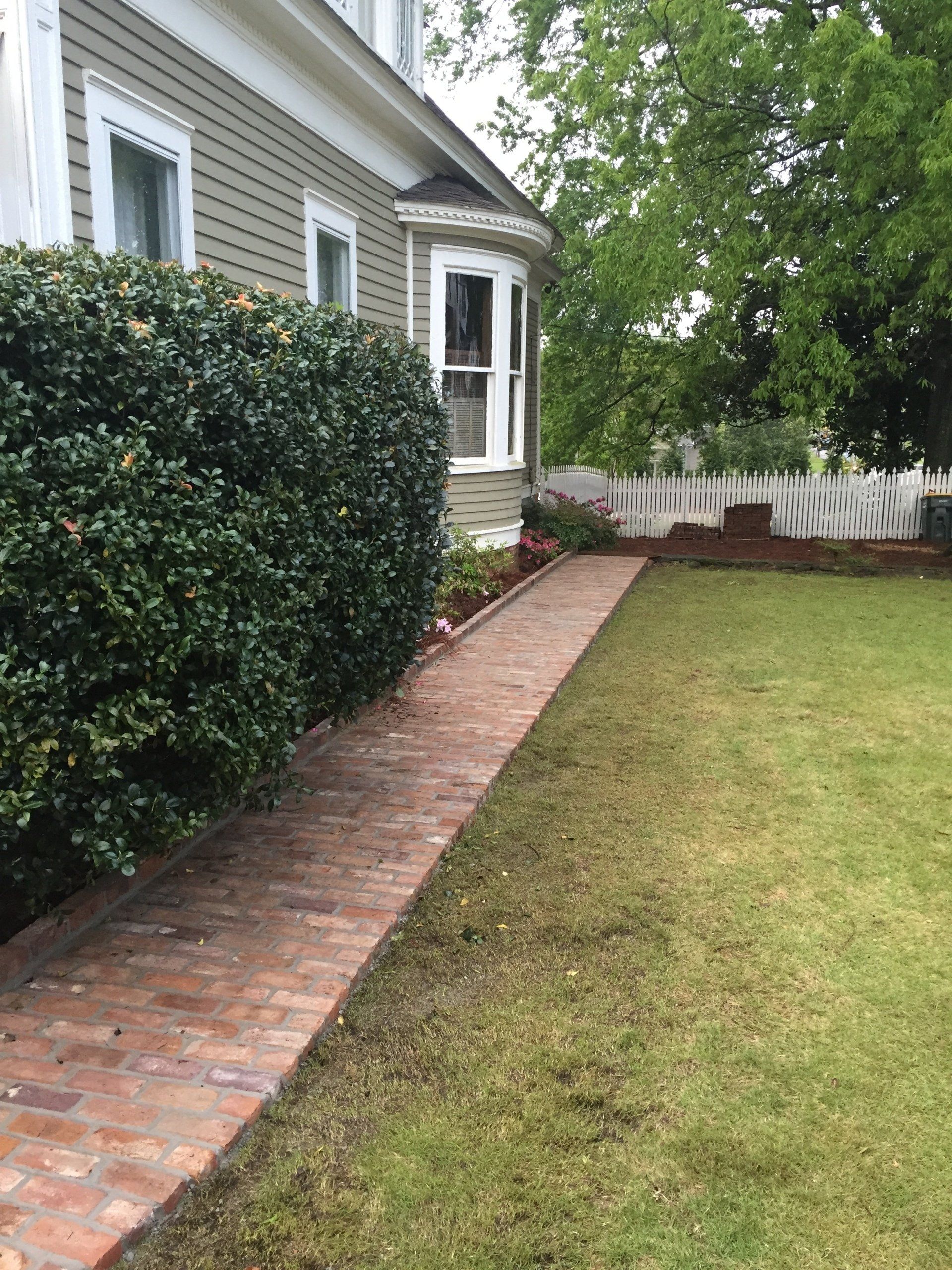 Brick path leads from lawn to house with a bay window, bordered by bushes and a white picket fence.