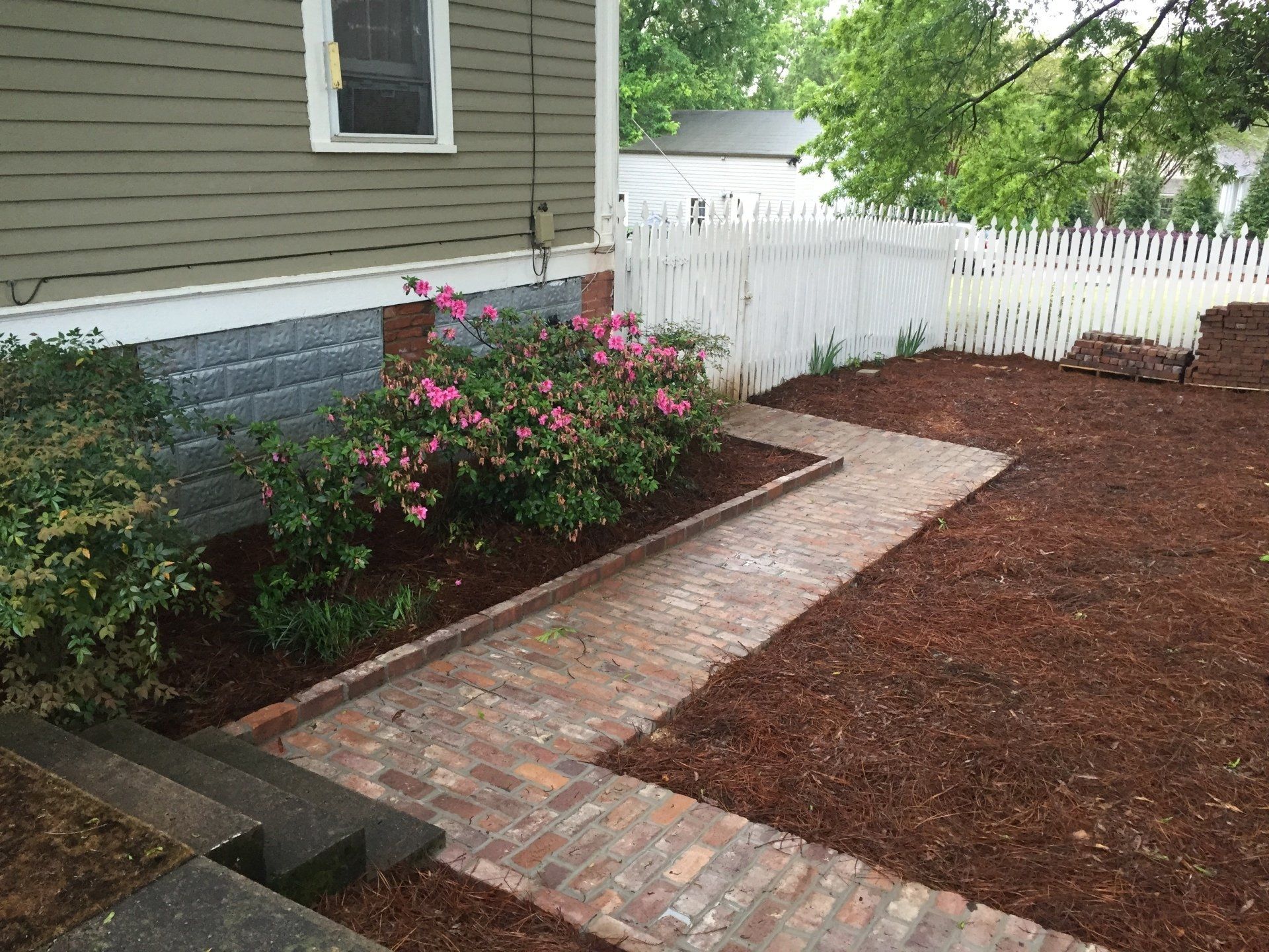 Brick pathway leads through a mulched yard with a picket fence, next to a house with shrubs and flowers.