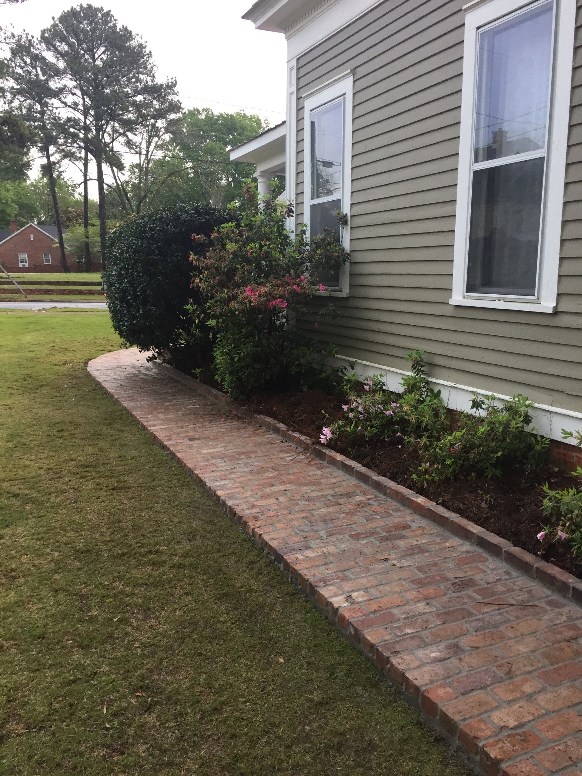 Brick walkway alongside a green house with windows and a landscaped garden bed.