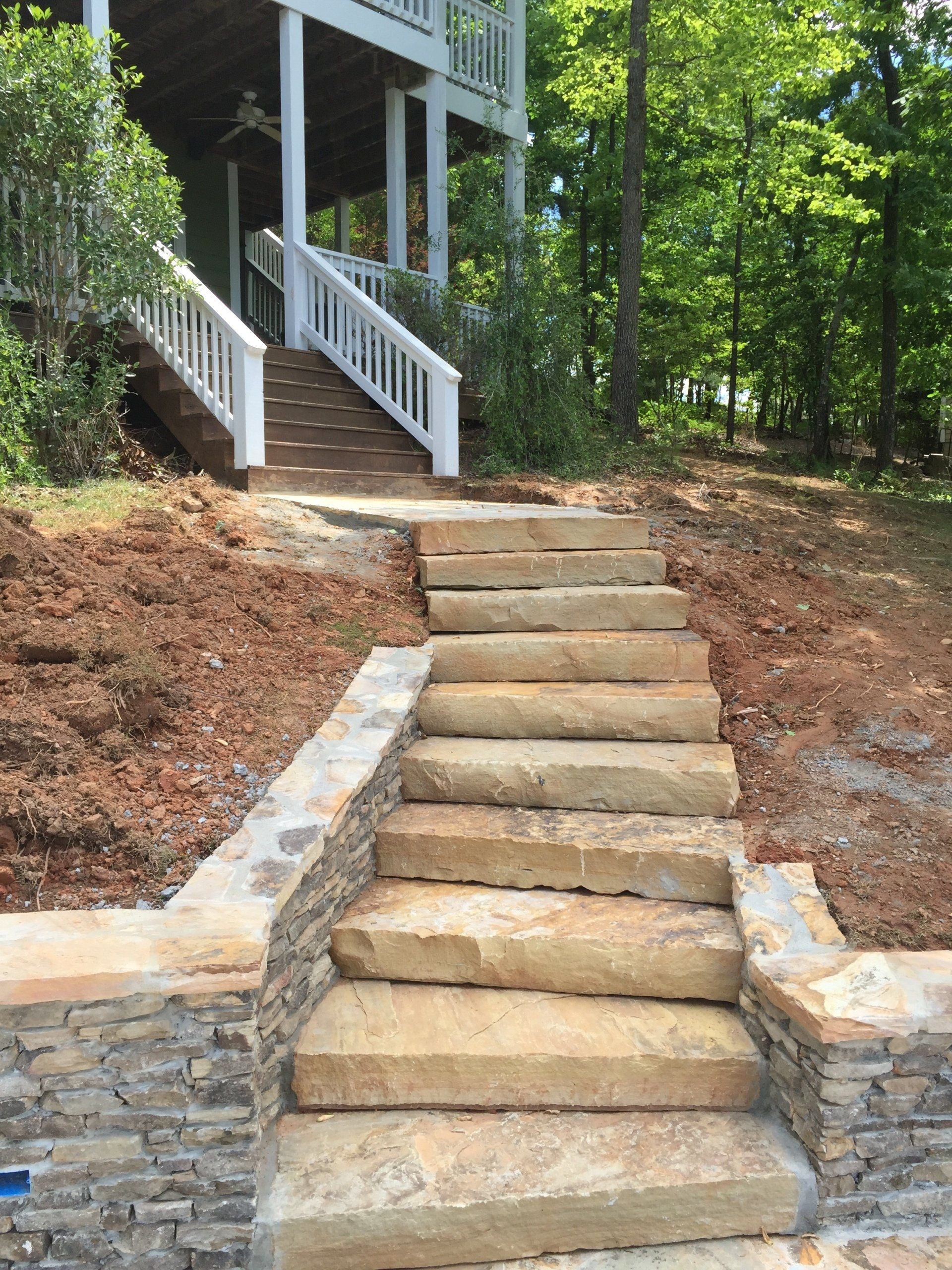 Stone steps leading up to a house with white railing, set in a wooded area with brown soil and lush green trees.