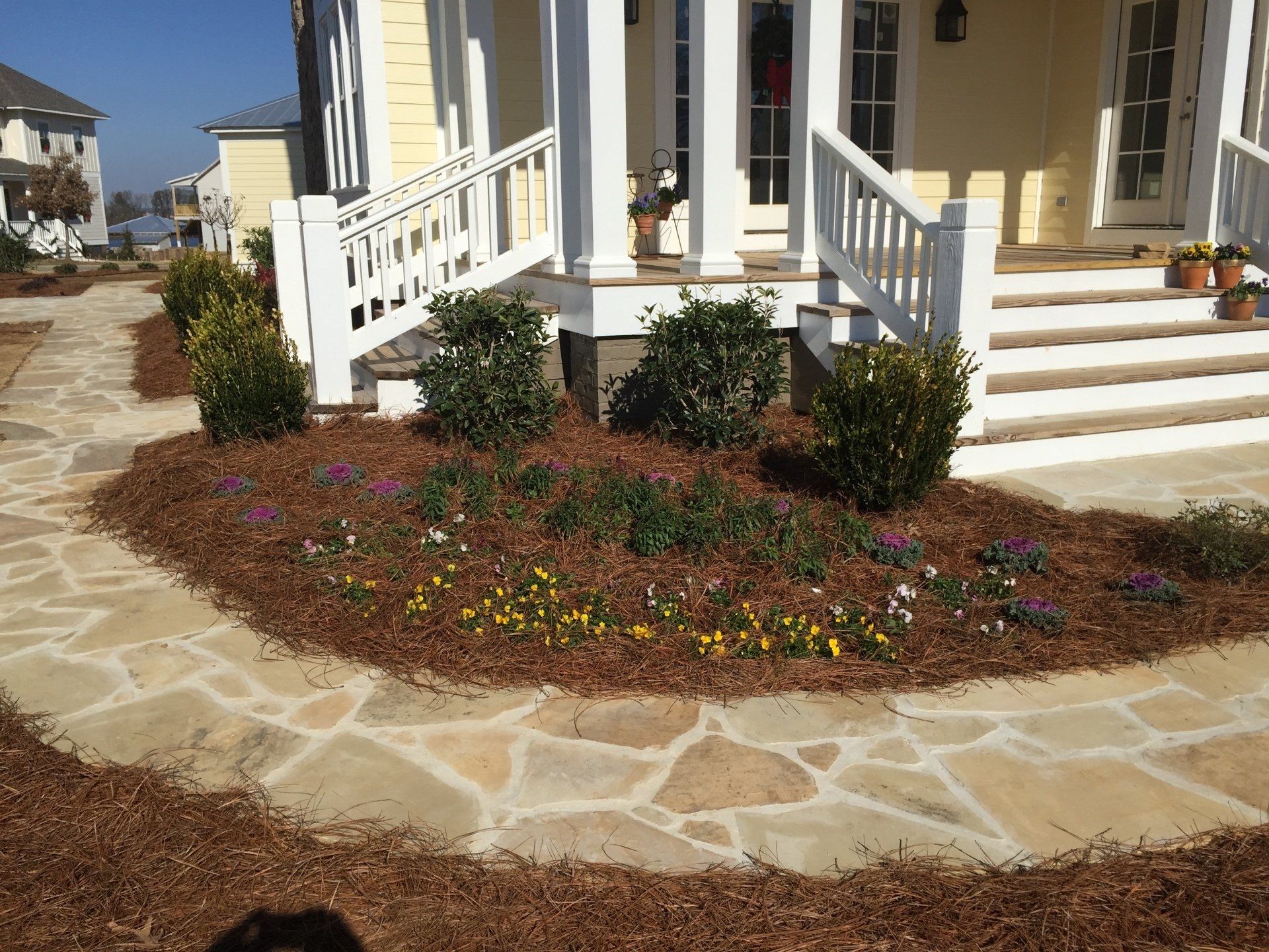 Stone walkway leads to a yellow house with a landscaped flower bed containing bushes and colorful flowers.