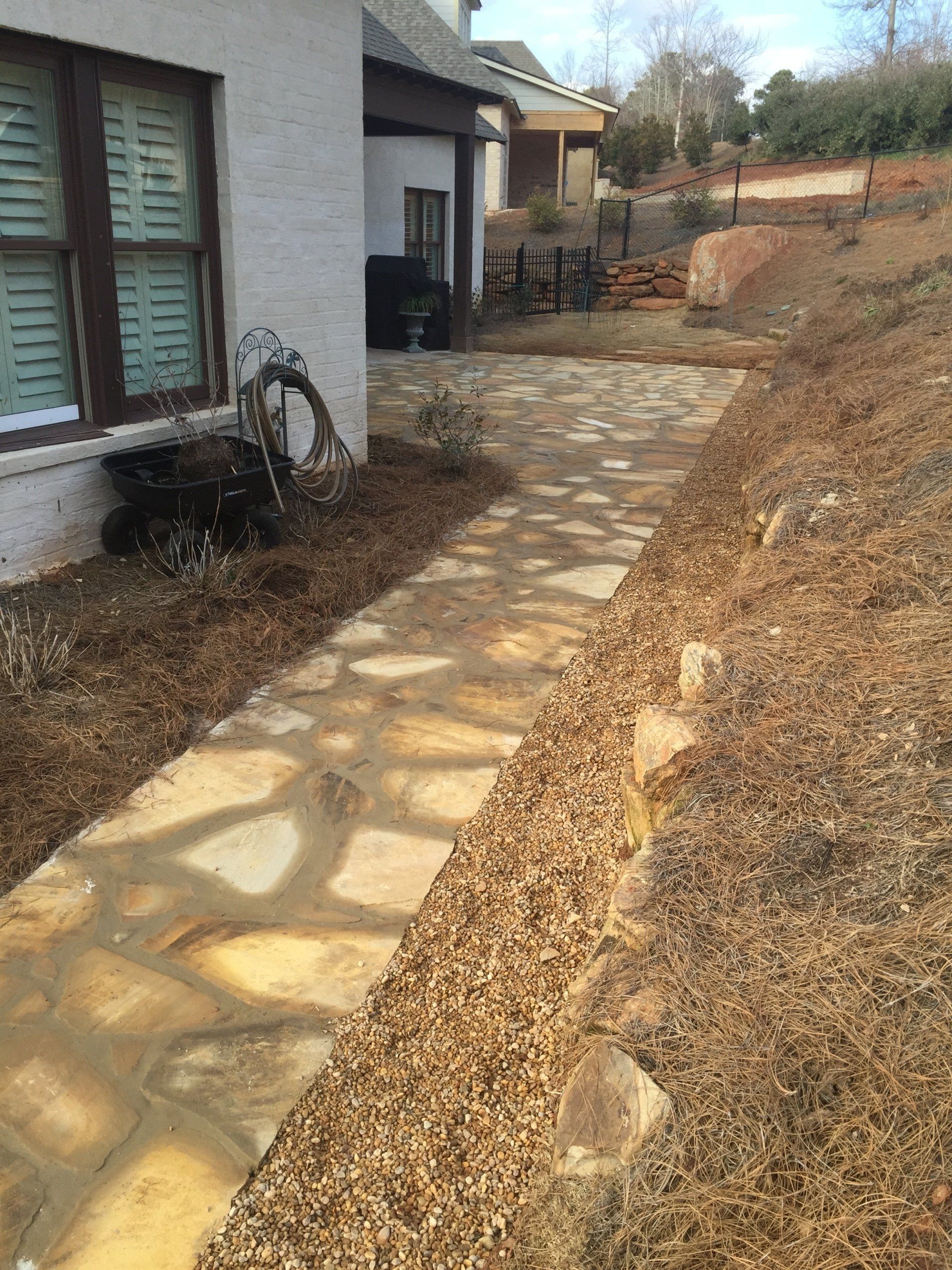 Flagstone walkway next to a beige house and sloped hillside, with brown pine needles.