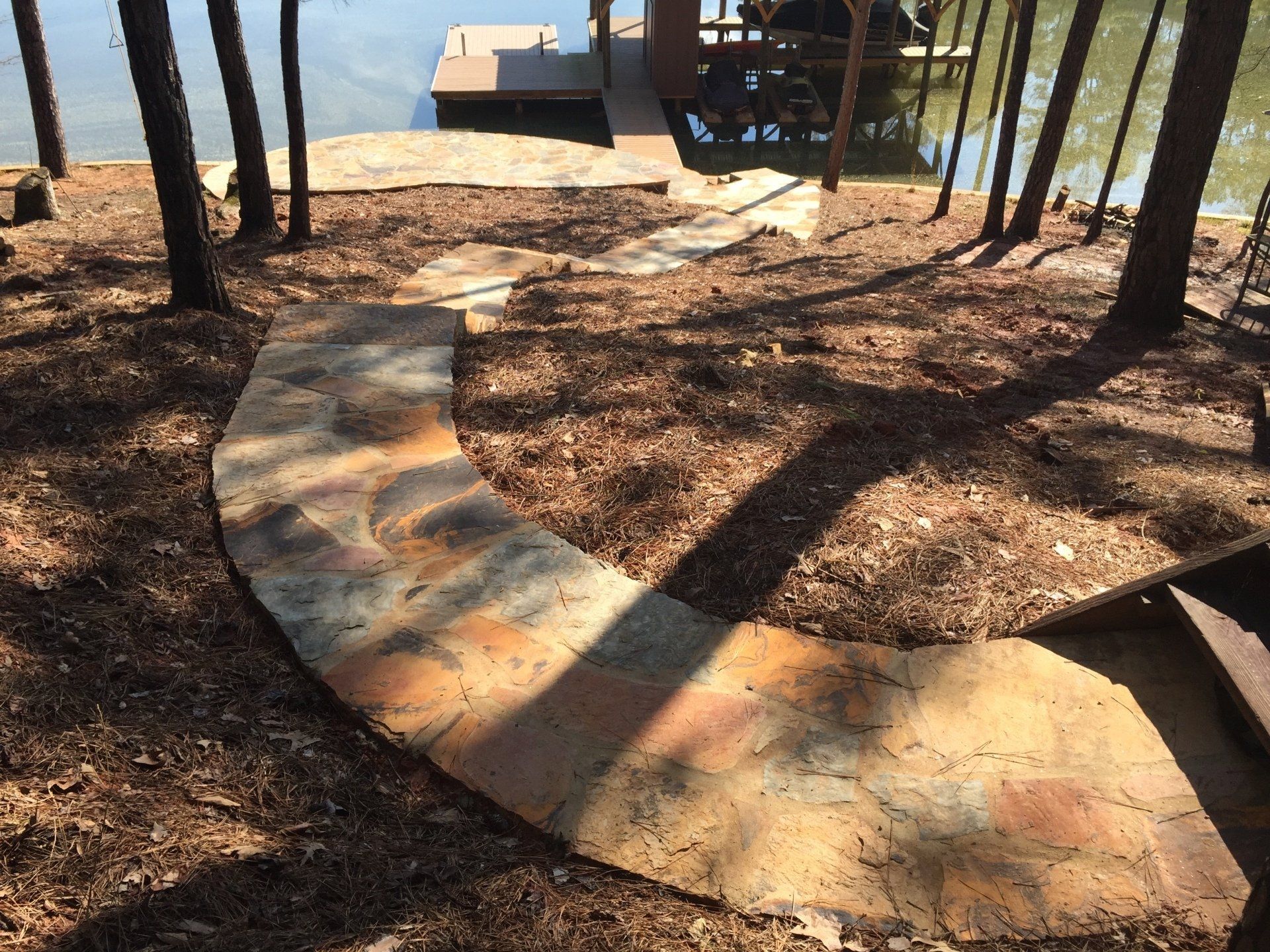 Stone pathway curves down to a dock on a lake, surrounded by trees and brown mulch.