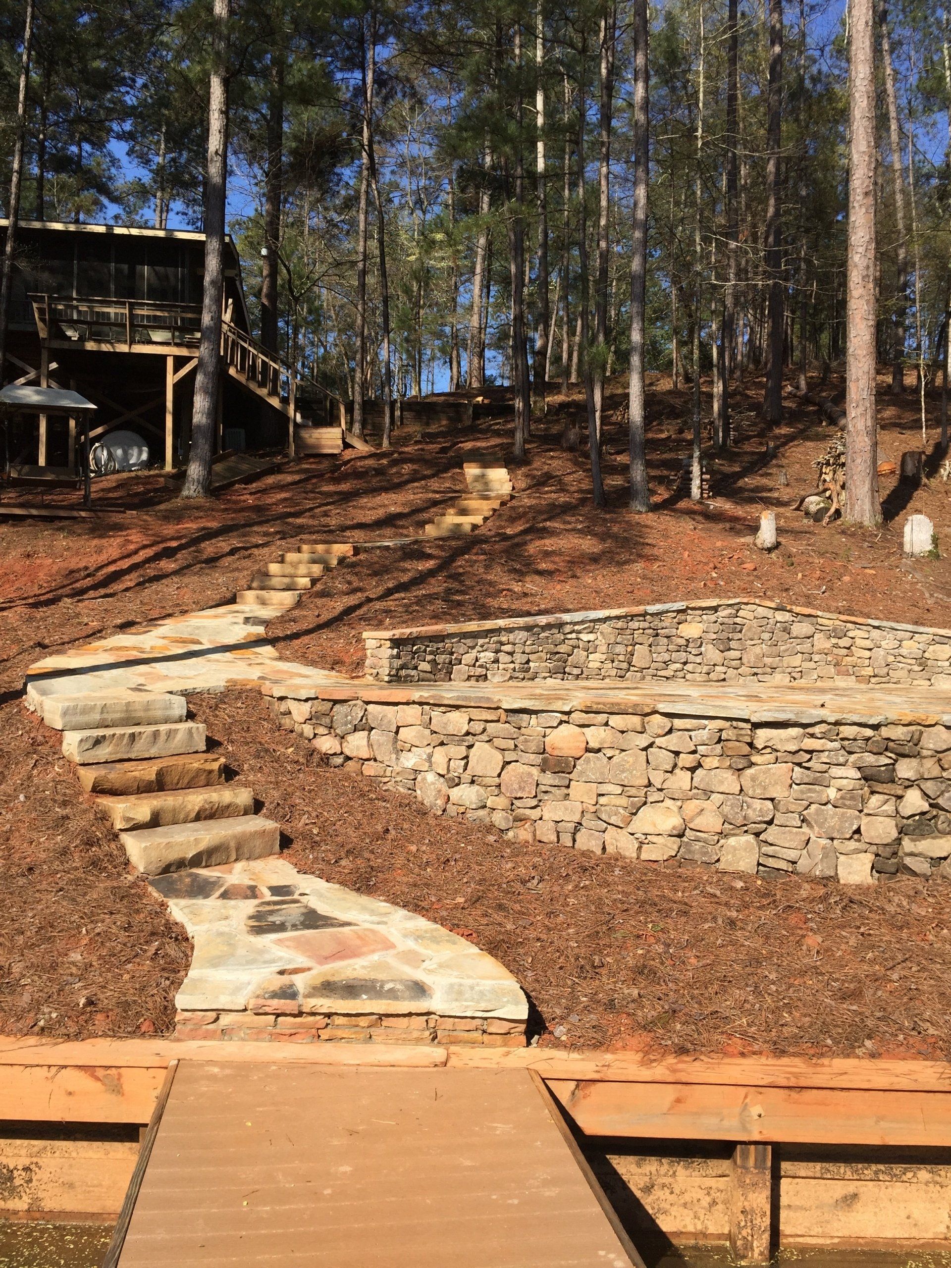 Stone steps and a pathway lead up a hill from a wooden dock to a stone retaining wall in a wooded area.