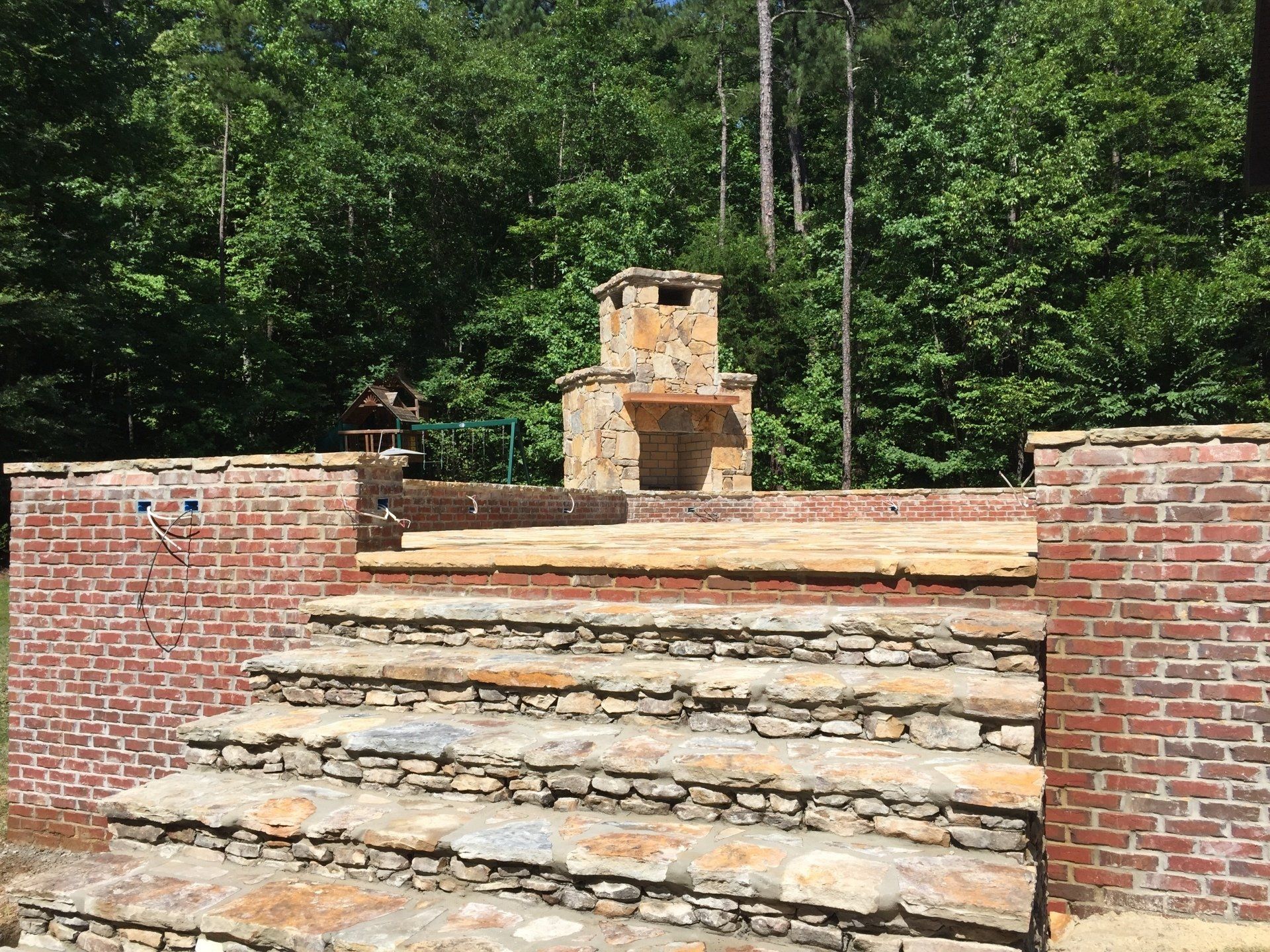 Stone steps leading to a brick and stone outdoor fireplace with a forest backdrop.