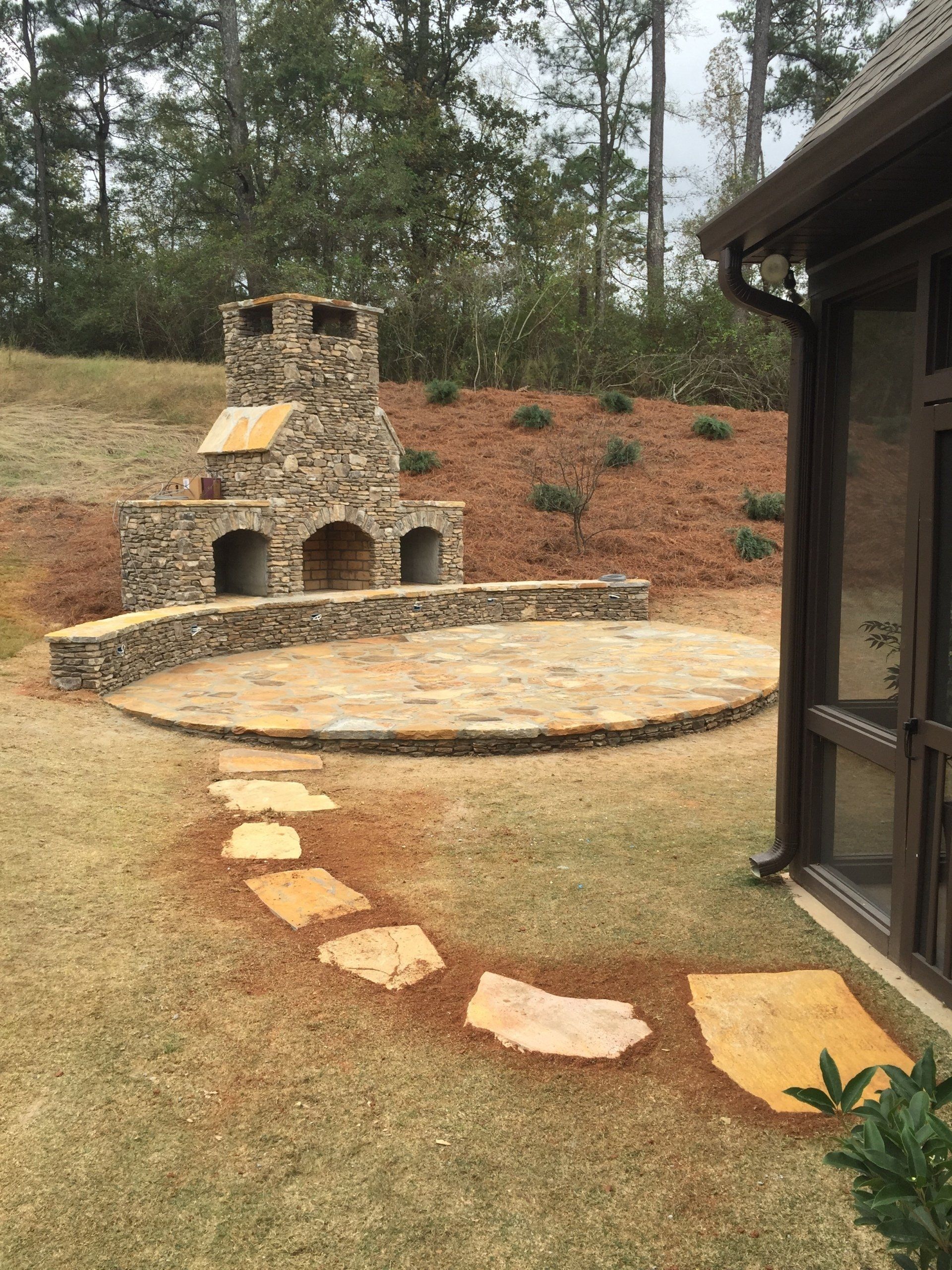Stone fireplace and patio in a backyard, pathway of stones leading to it.