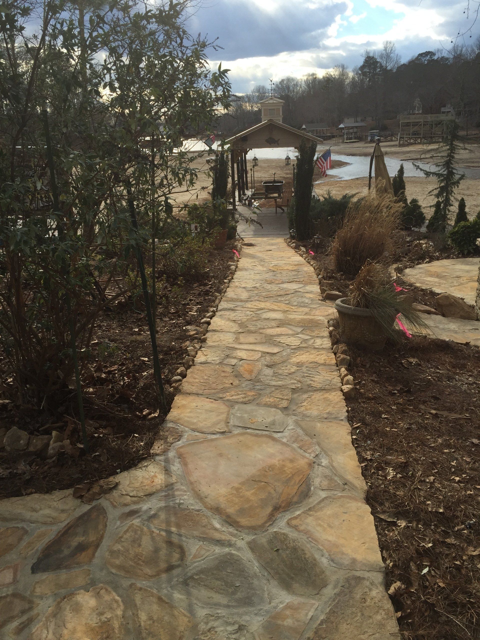 Stone path leading to an archway overlooking a lake. Lush greenery on either side. Cloudy sky.