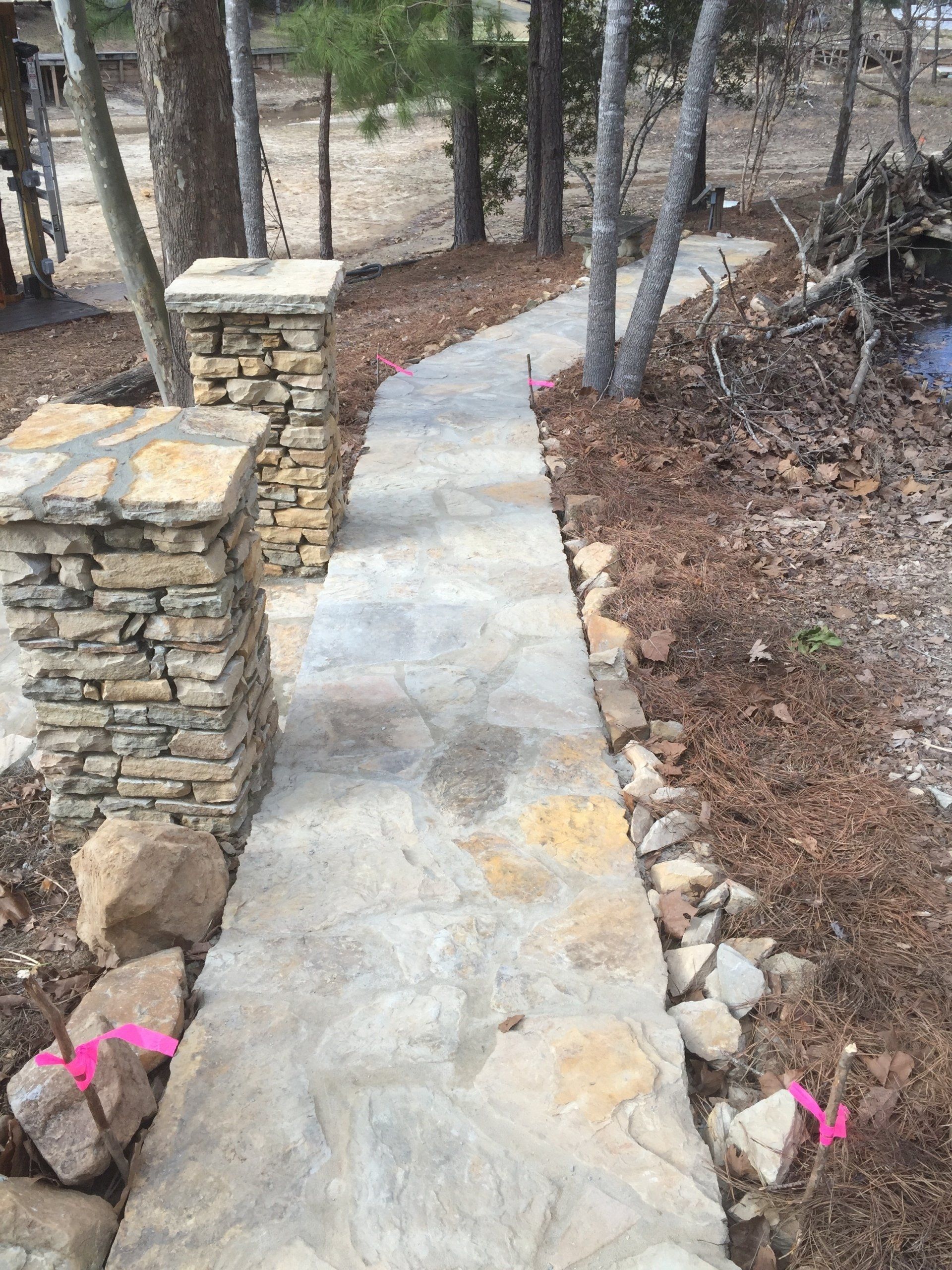 Stone path leads through wooded area, flanked by stone pillars and small rocks.