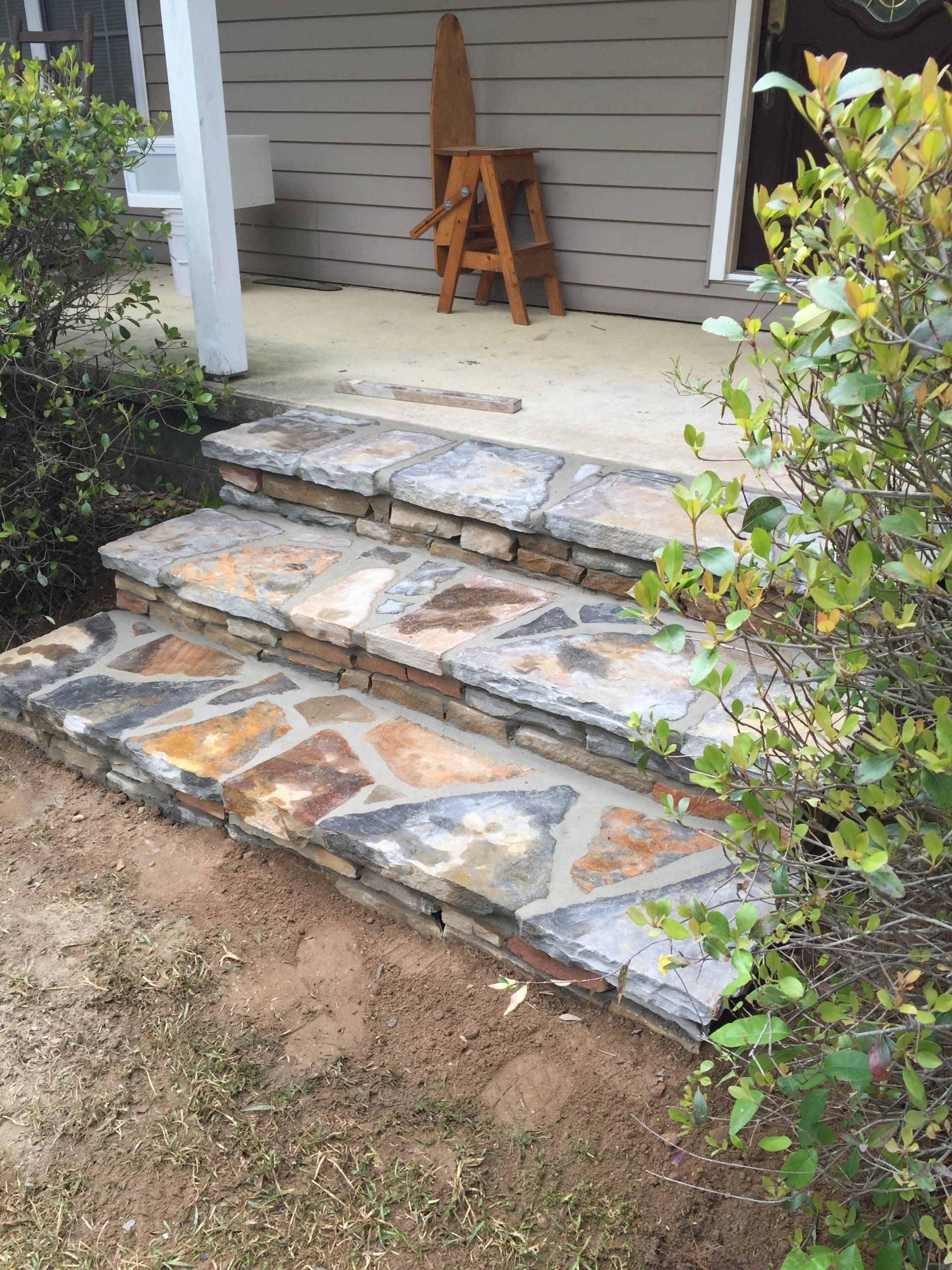 Stone steps leading up to a porch. Tan, gray, and brown stones. Wooden chair on porch.