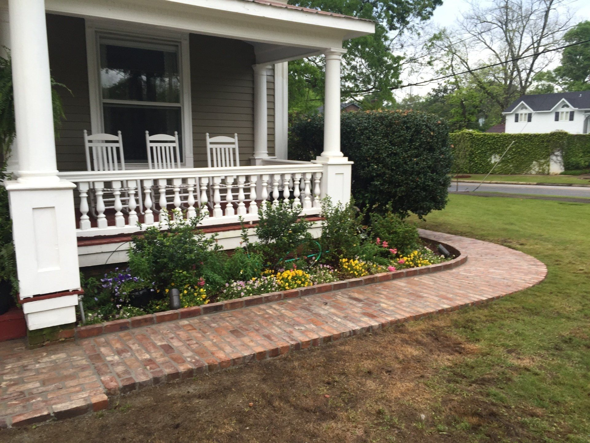 Brick path curving to a porch with white columns, a garden of flowers, and a house.