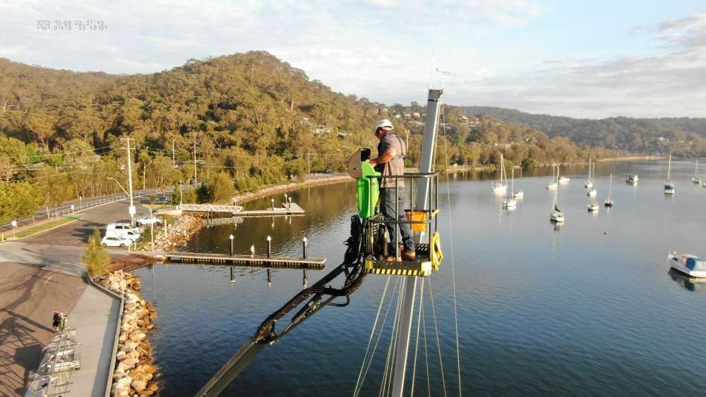 A Man Is Standing On A Crane Overlooking A Body Of Water — High Work Services in Maitland, NSW