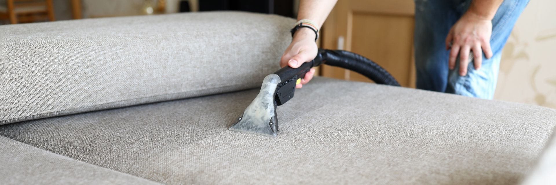 A person uses a handheld vacuum upholstery cleaning tool to clean a light gray fabric sofa.