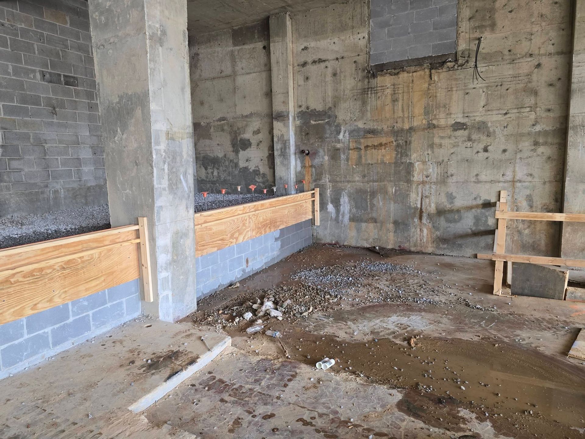 Abandoned unfinished concrete room with cinder block walls, muddy floor, and wooden shelving unit